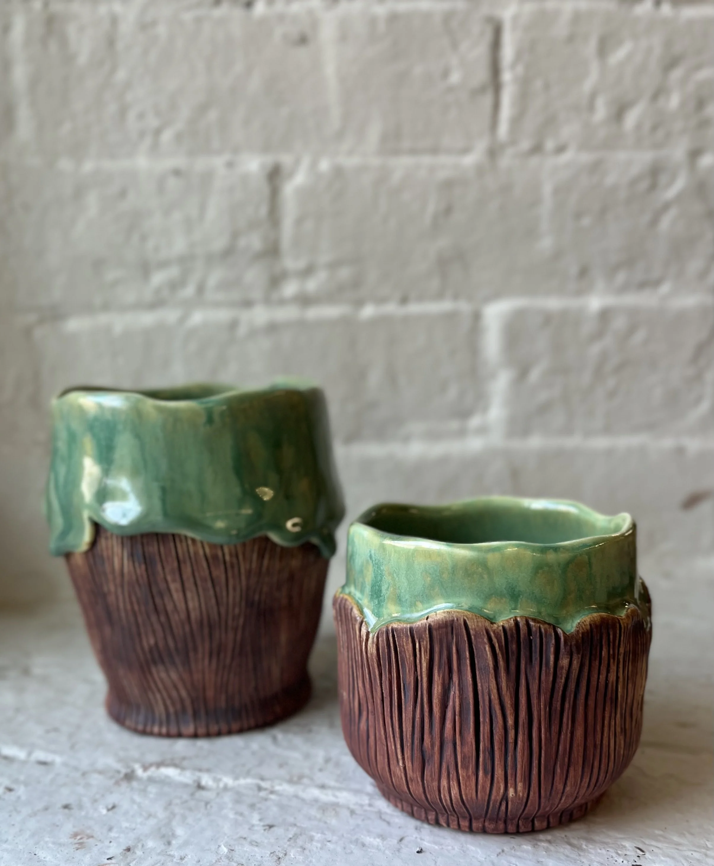 Two ceramic cups with textured brown base and green glazed top trim, placed on a white surface against a white brick wall background.