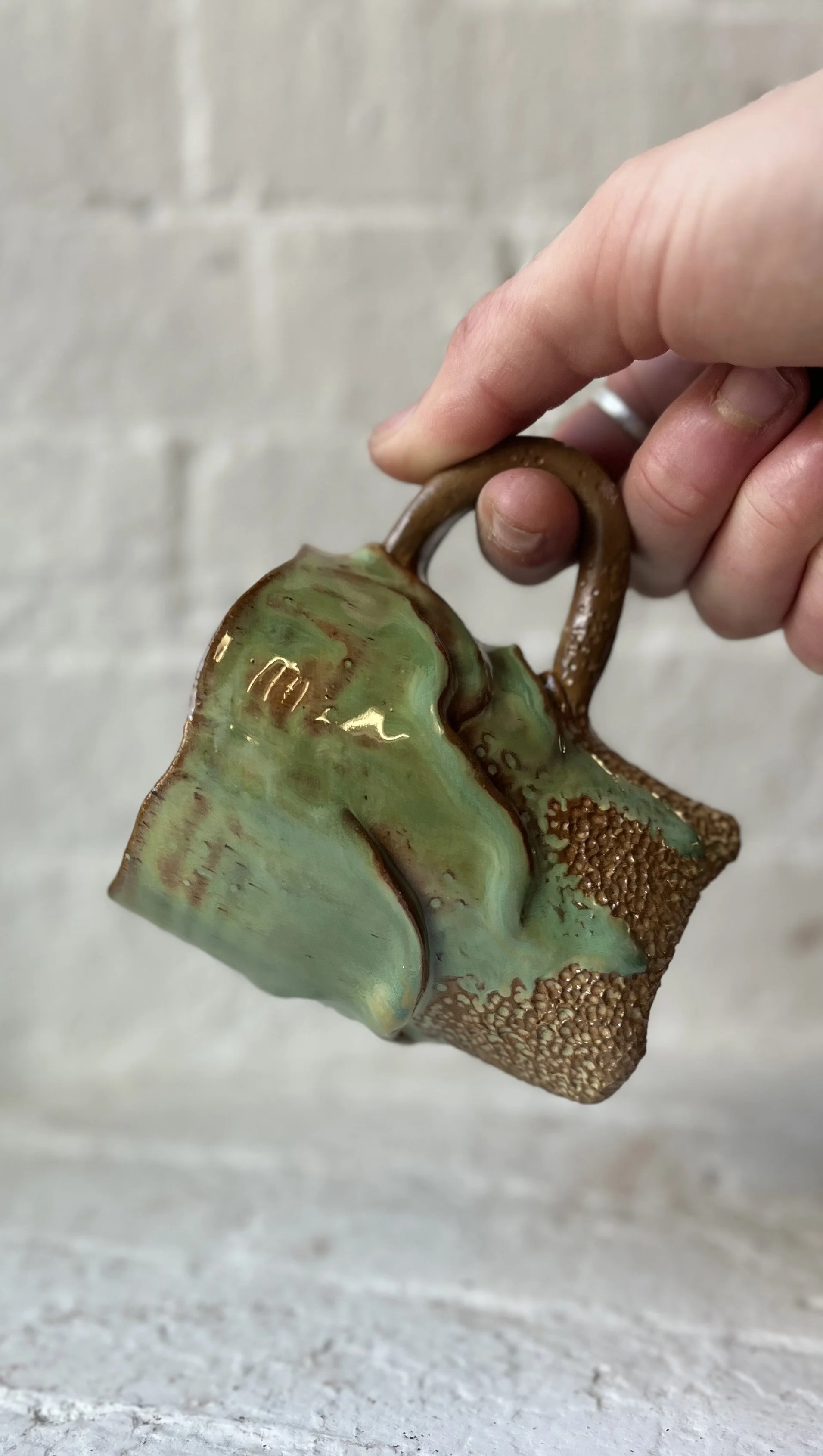 Person holding a small glass jar with a handle, featuring green and brown swirled glass art.