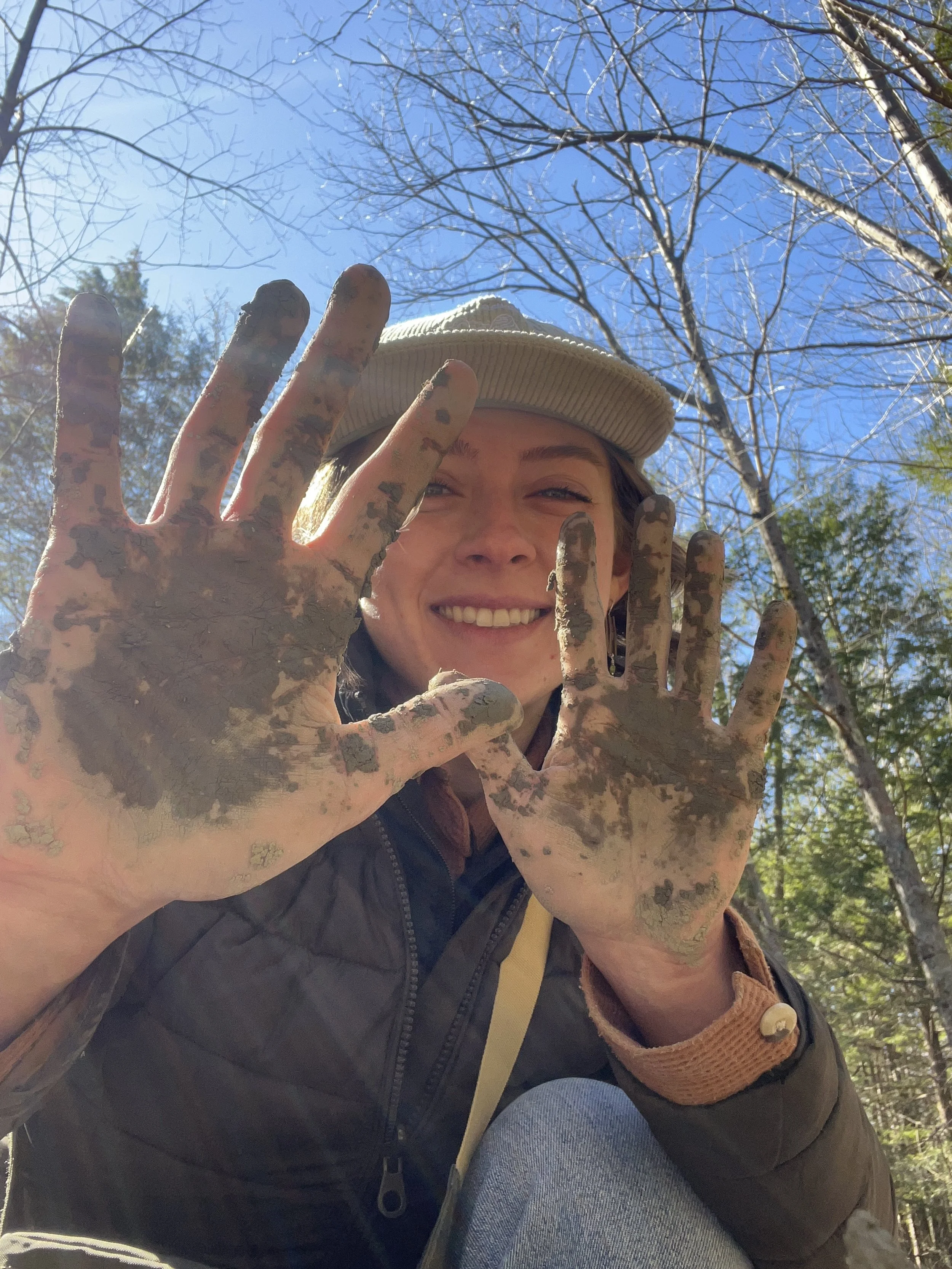 Young woman smiling outdoors on a sunny day, showing hands covered in mud, with leafless trees and blue sky in the background.