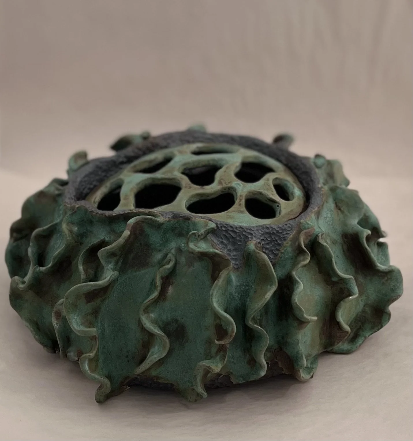 Ceramic art piece resembling a sunflower seed with textured petals and a lattice pattern on top, in shades of green and black.