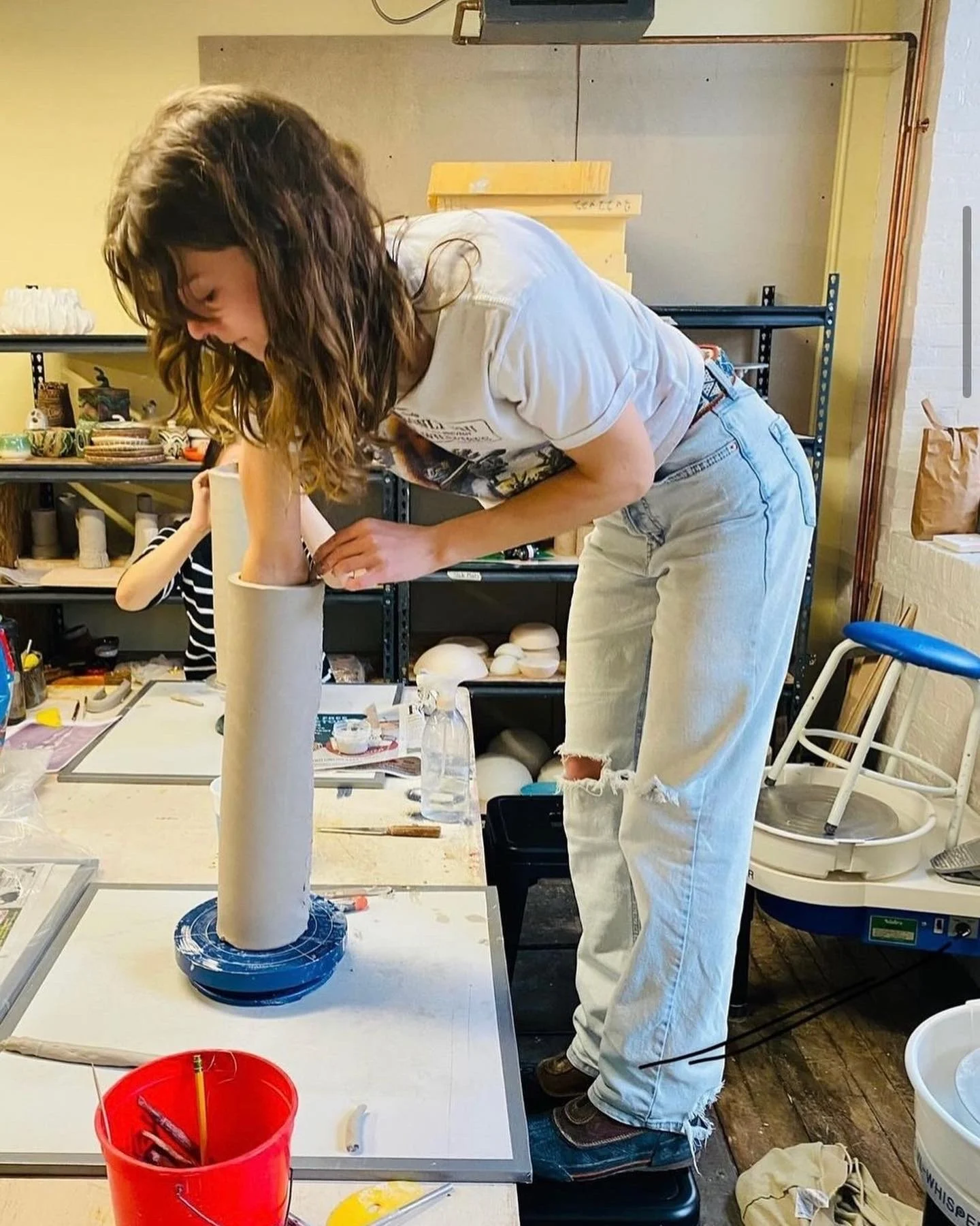 A woman with long, wavy brown hair, wearing a white T-shirt and ripped jeans, is working on a large cylindrical sculpture in an art studio. She is leaning over the sculpture, which is placed on a flat surface, and appears to be shaping or assembling 