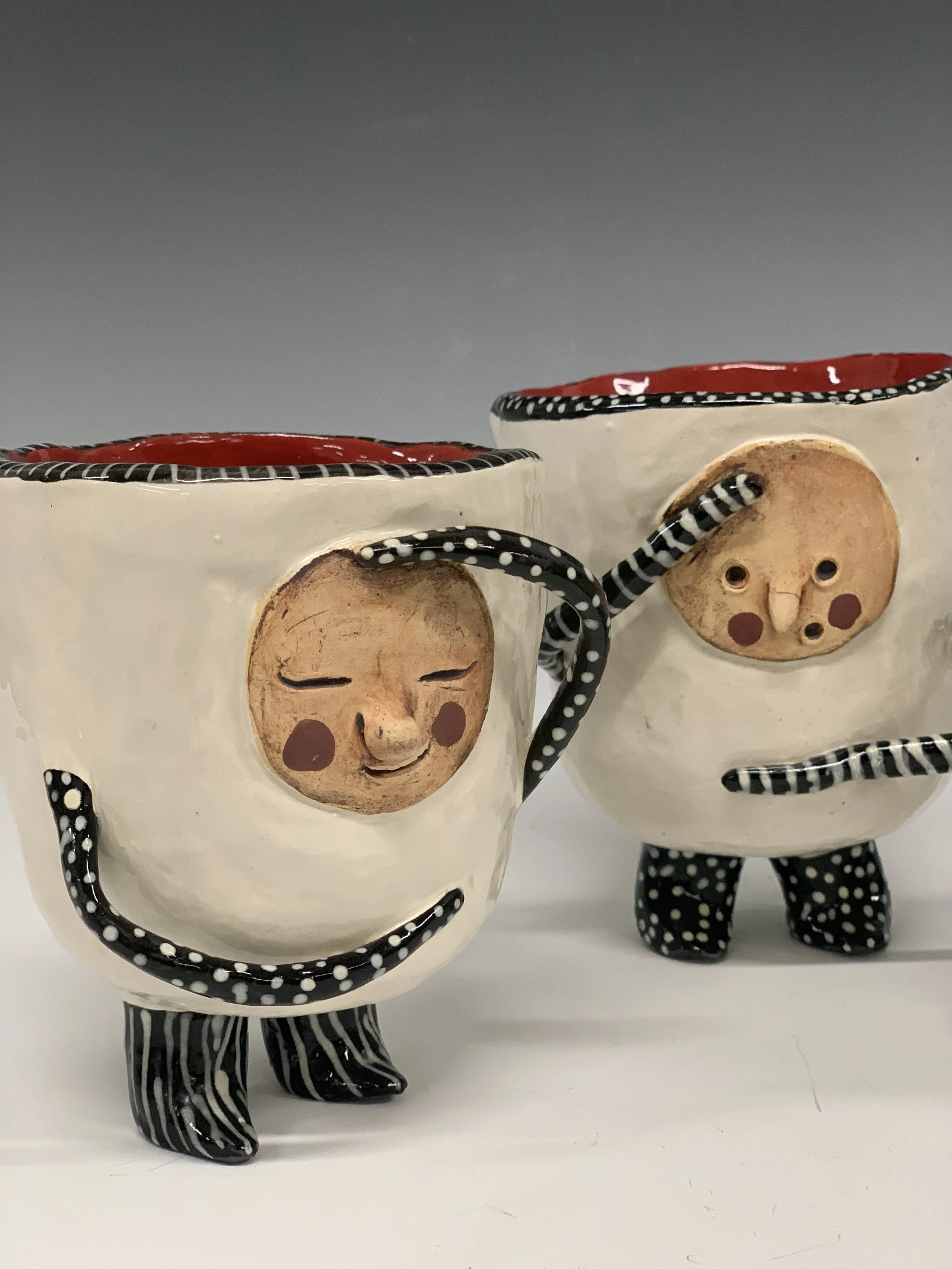 Two ceramic mugs with whimsical face designs, striped legs, and polka-dotted handles, positioned on a white surface against a gray background.