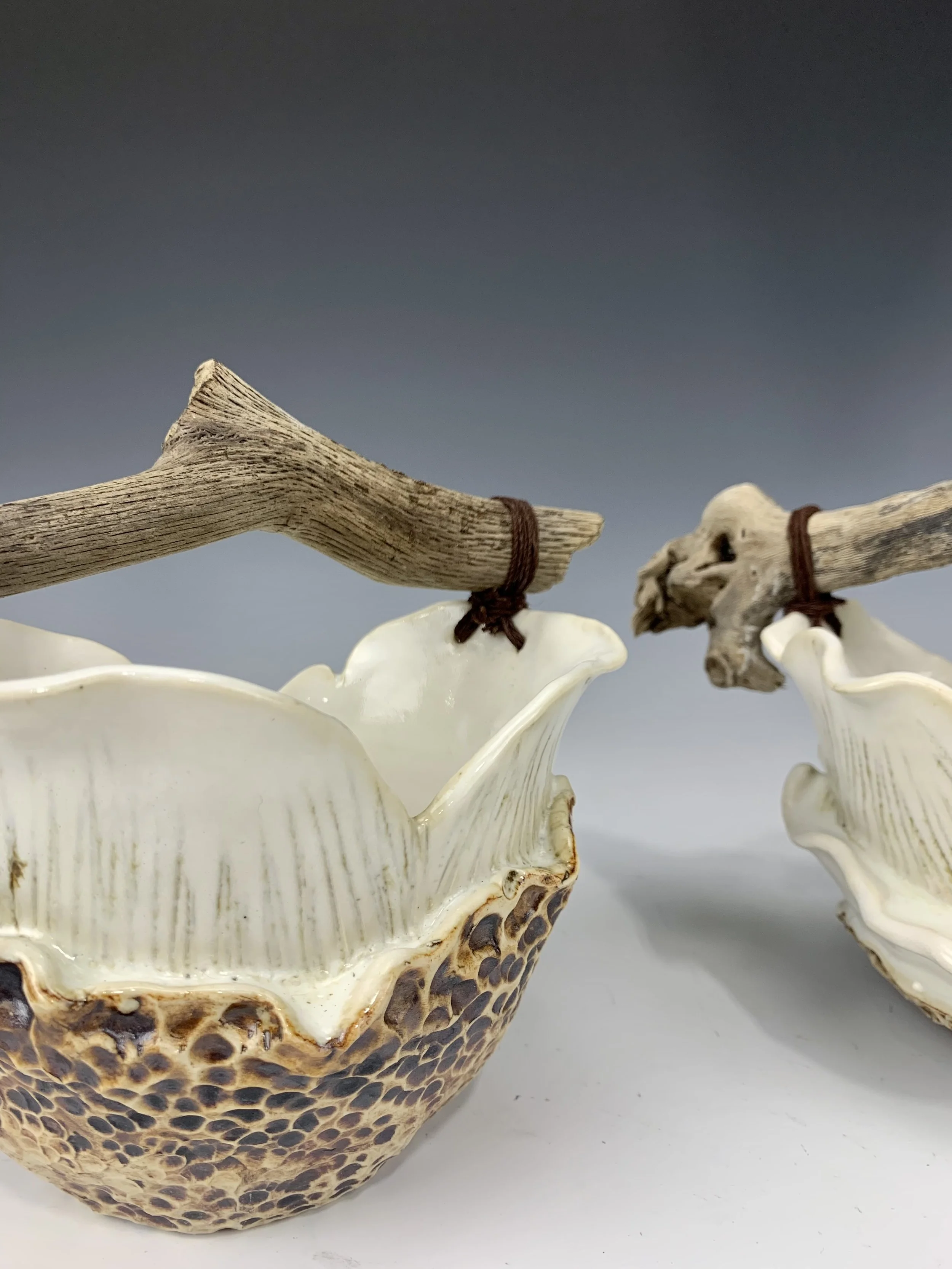 Two ceramic bowls with a textured, brown exterior resembling a shell pattern, and cream-colored interior, connected by driftwood tied with brown string.