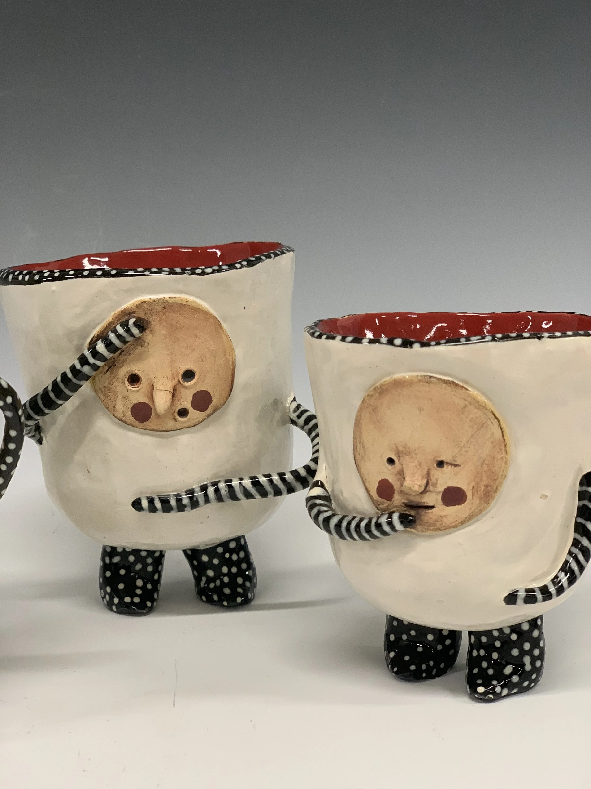 Two decorated ceramic mugs with faces, polka dot limbs and legs, and red interior, resembling whimsical characters.