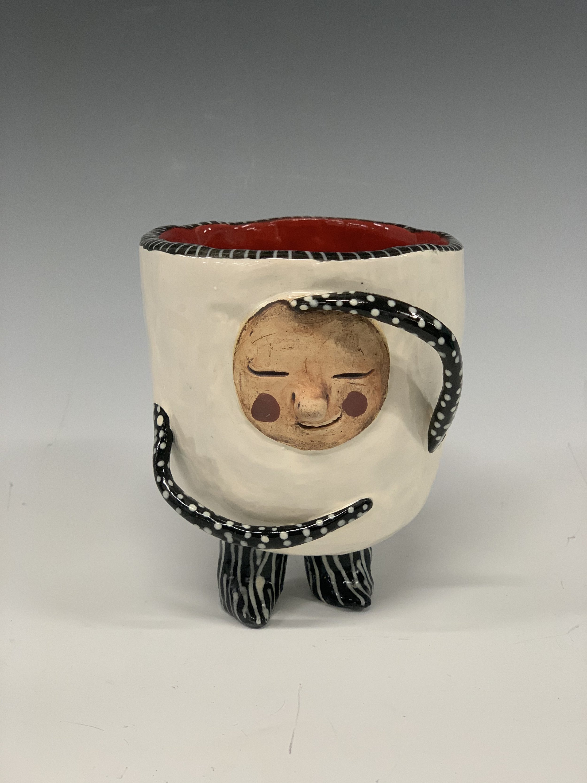 A ceramic mug with an anthropomorphic face and black and white striped arms and legs. The mug is white with a red interior and a black rim. The face has closed eyes, red cheeks, and a small smile.