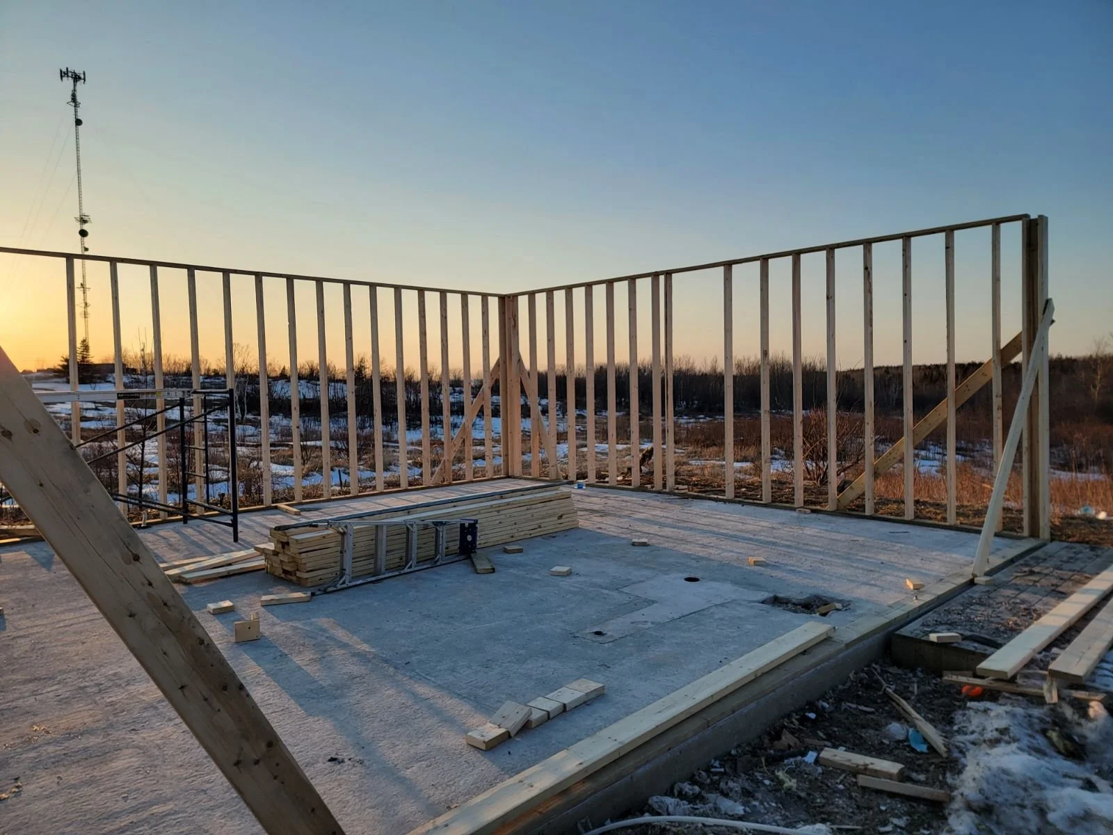 Construction site with wooden framing for a building, some stacked wood and tools on a concrete slab, and a snowy landscape in the background at sunset.