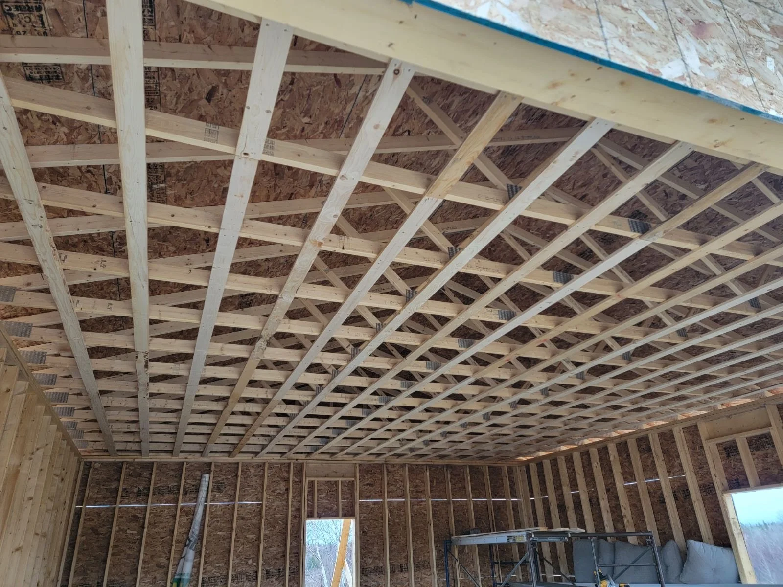 Interior view of a room under construction with wooden framing on the ceiling and walls, windows without glass installed, and construction tools and materials present.