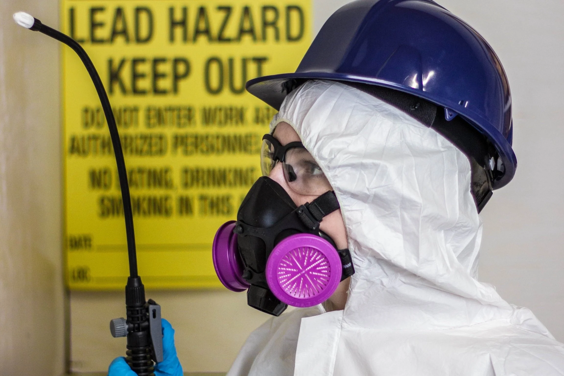 A worker dressed in protective gear, including a white hazmat suit, purple respirator mask, safety goggles, a blue hard hat, and blue gloves, holding a spray or inspection tool. In the background, a yellow warning sign reads 'Lead Hazard Keep Out'.