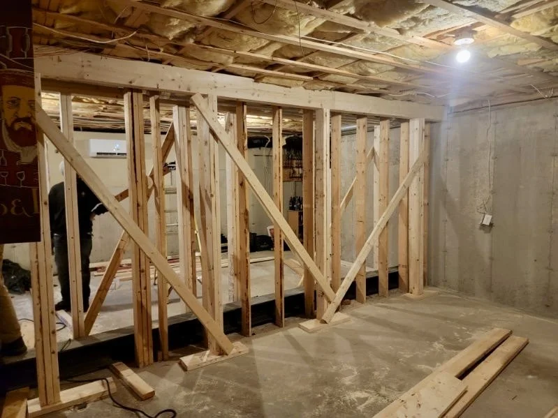 Indoor construction site with wooden framing for walls, concrete floor, unfinished ceiling with insulation, and a single light fixture.