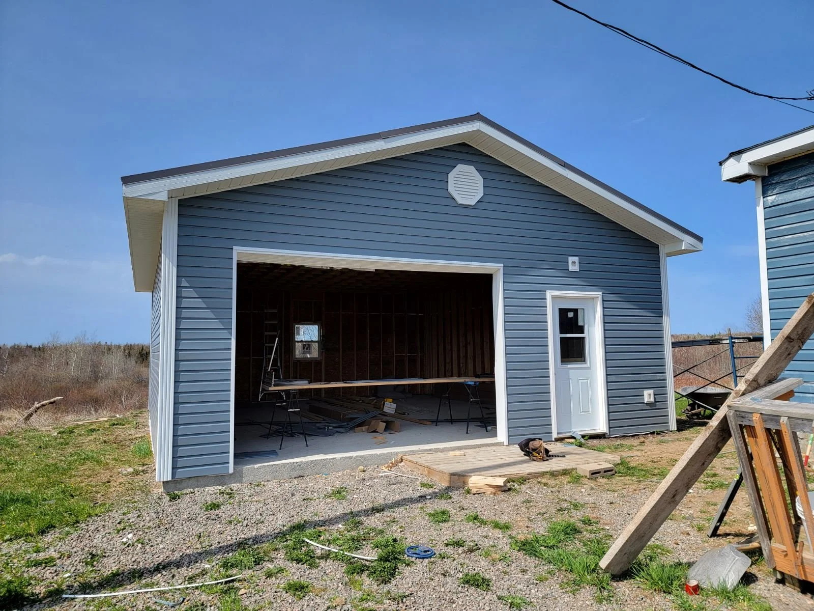 A small blue garage with a white door and trim under construction. The garage has vinyl siding and is situated on a gravel and dirt lot, with construction materials inside and outside.