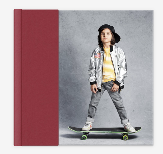 Child standing on a skateboard indoors, wearing a silver jacket, striped shirt, jeans, and a backward cap, next to a maroon border.