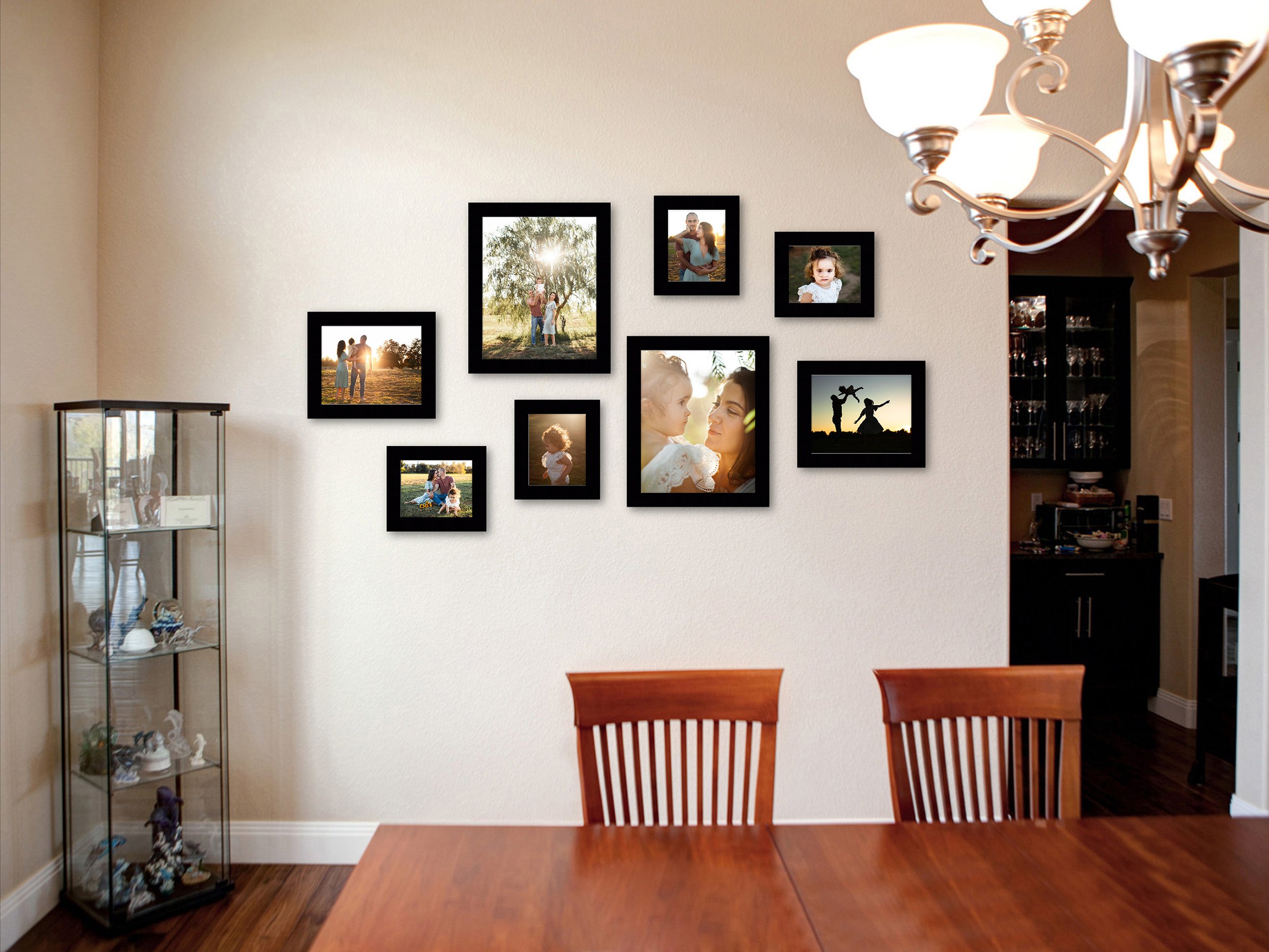Dining room with a gallery wall of family photos in black frames, a chandelier, wooden dining table and chairs, a glass cabinet with decorative items, and a kitchen background.
