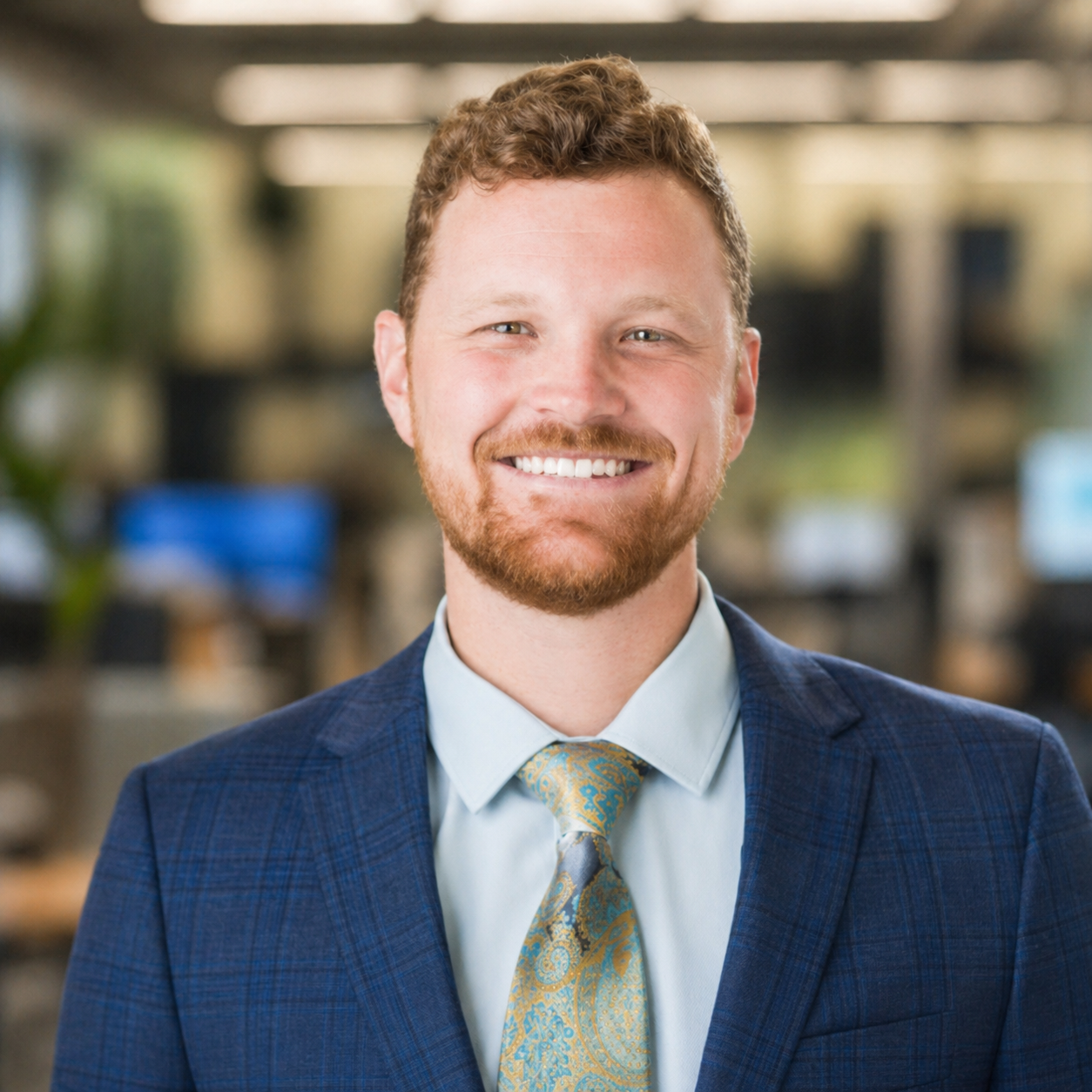A smiling man with curly red hair and a beard, wearing a blue plaid suit, light blue shirt, and a patterned yellow and blue tie, standing in a blurred office environment.