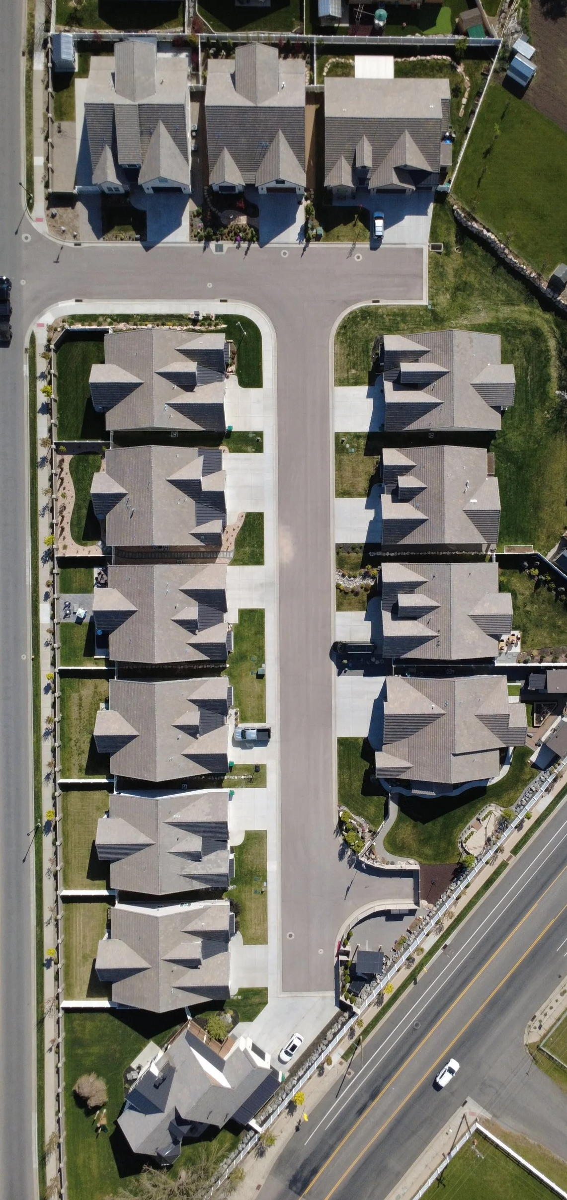 An aerial view of a suburban neighborhood showing several houses with grey roofs, driveways, and green lawns, along with a street at the bottom.