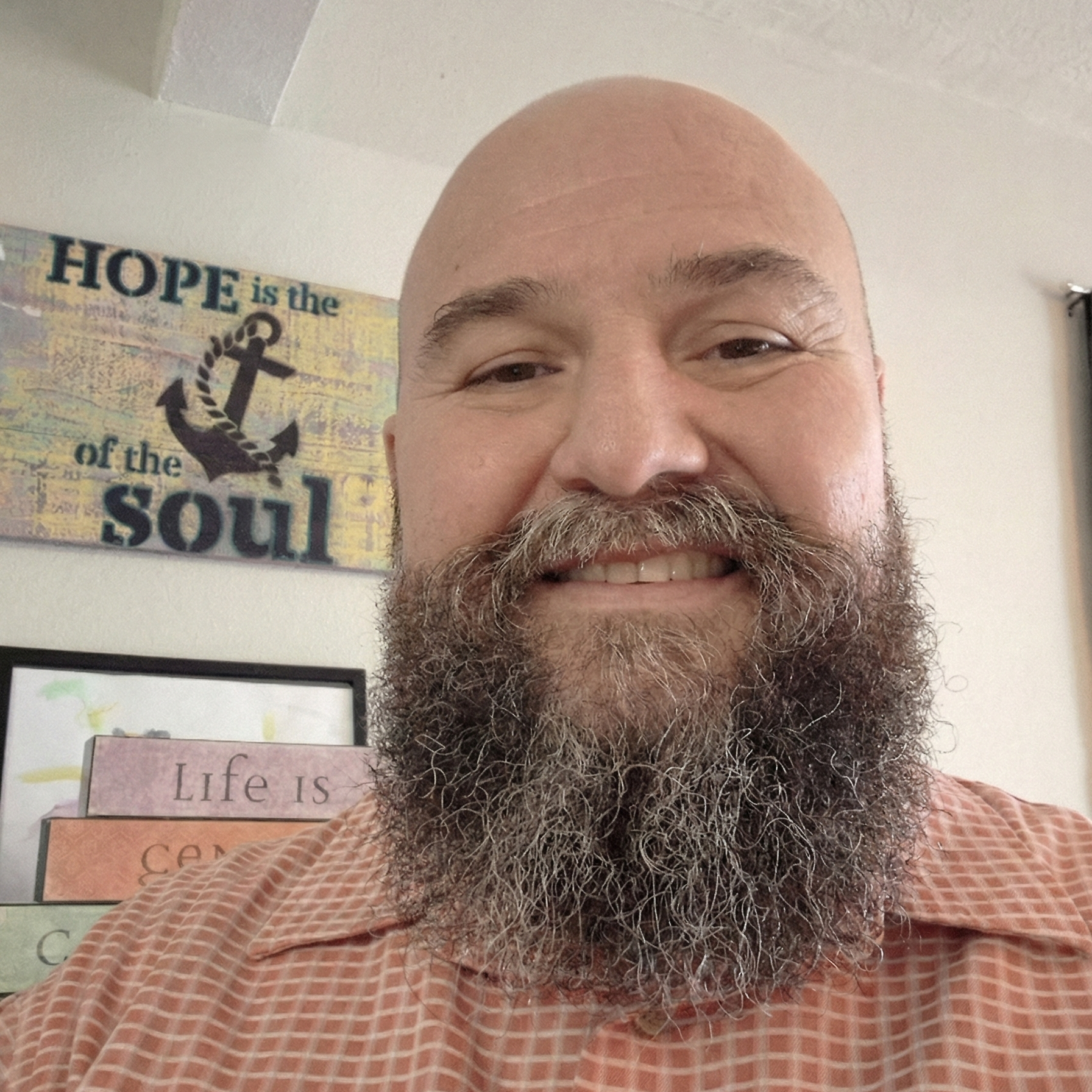 A smiling man with a bald head and a full beard, wearing a checkered shirt, in front of wall art that reads 'HOPE is the anchor of the soul' and 'Life is...'