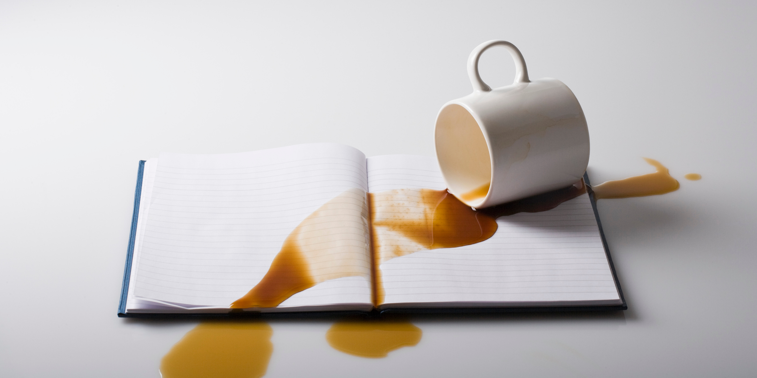 Image of while coffee mug on top of an open notebook with dark brown coffee spilling out of the mug and onto the blank notebook