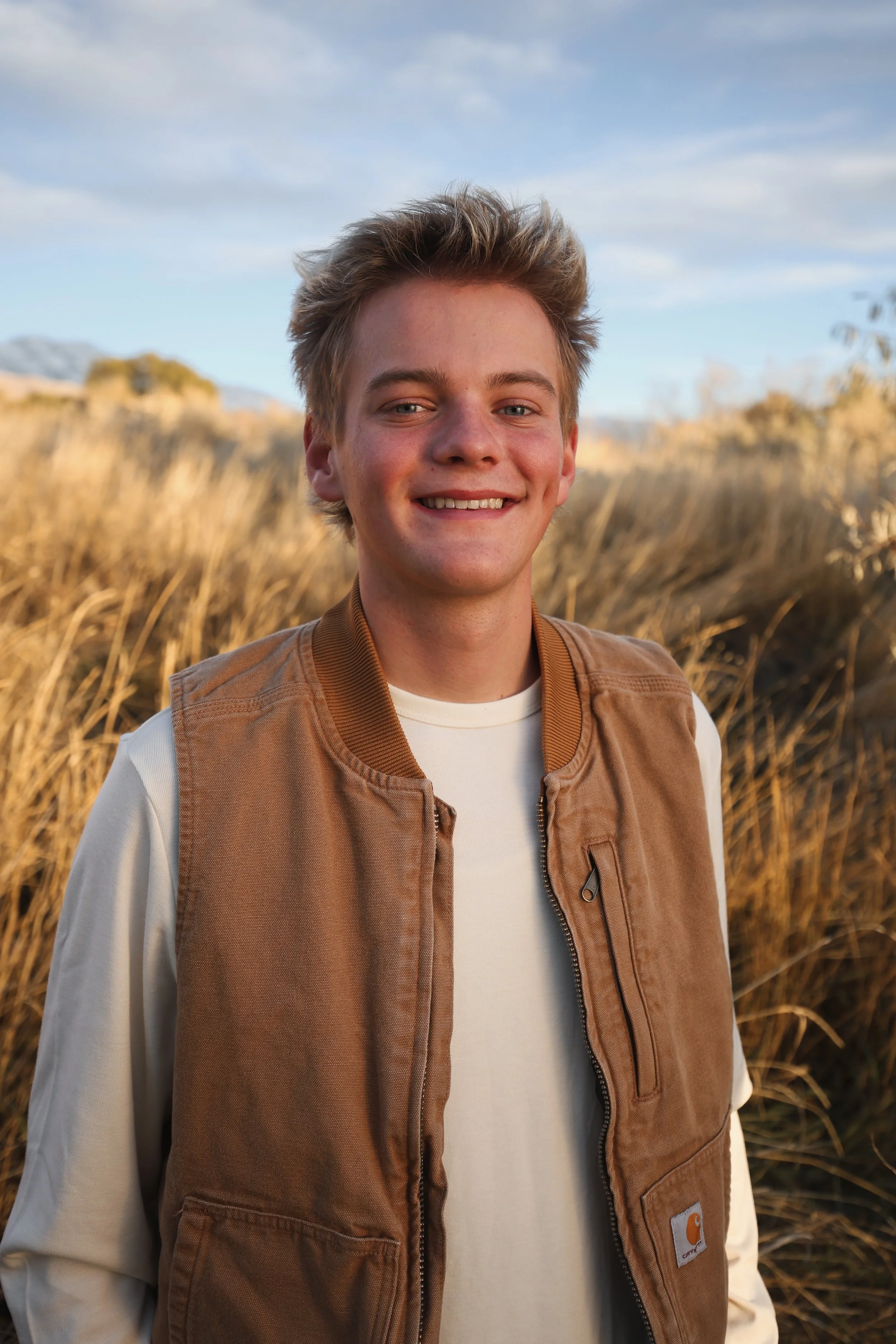 Smiling young man with blonde hair wearing a beige Carhartt vest and off-white shirt standing outdoors in a field of tall, golden grasses with mountains in the background.