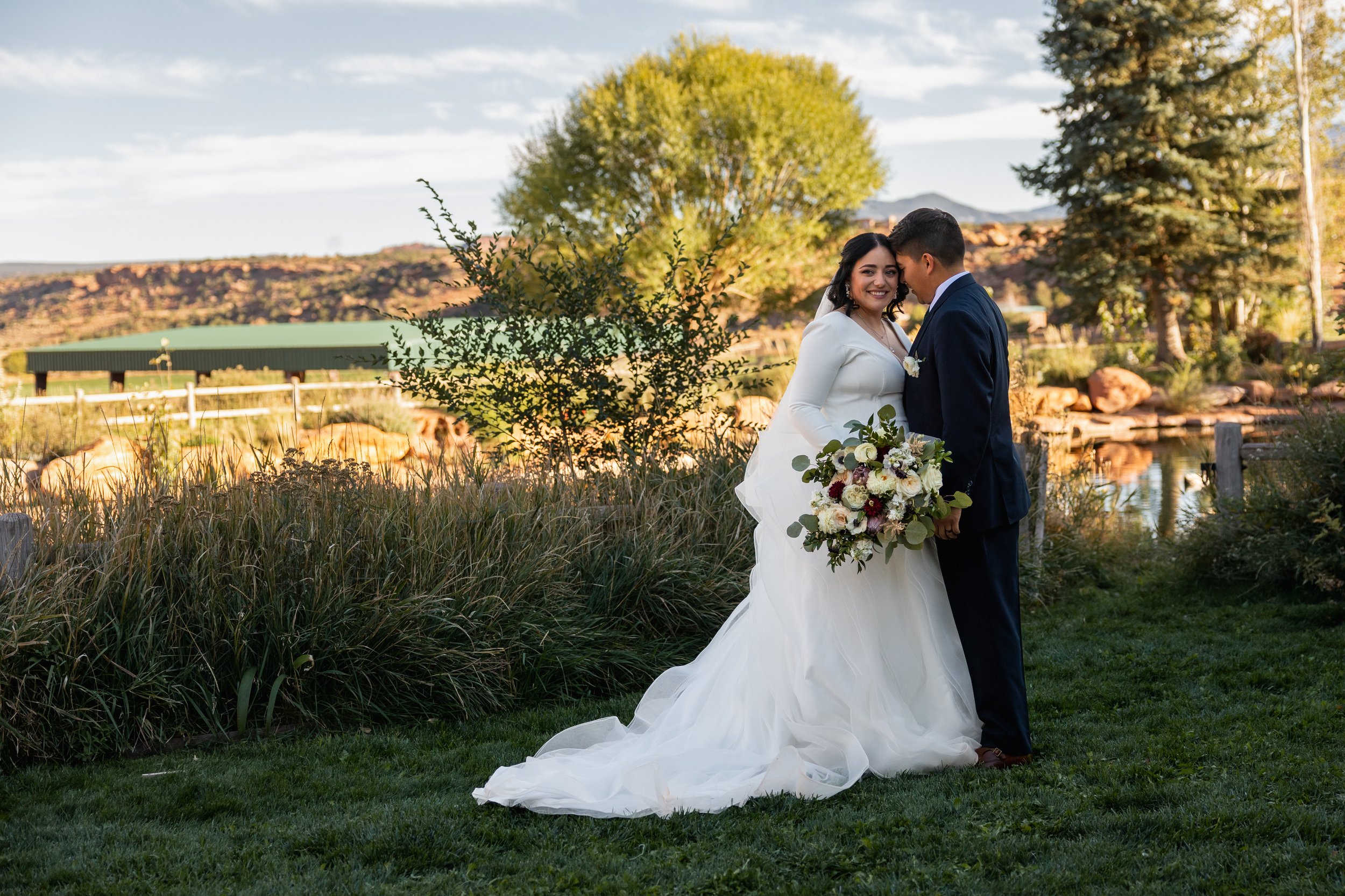 A bride and groom are standing together outdoors near a pond, with trees and mountains in the background during sunset. The bride is in a white wedding gown holding a bouquet, and the groom is in a navy suit. They are smiling and looking at each othe