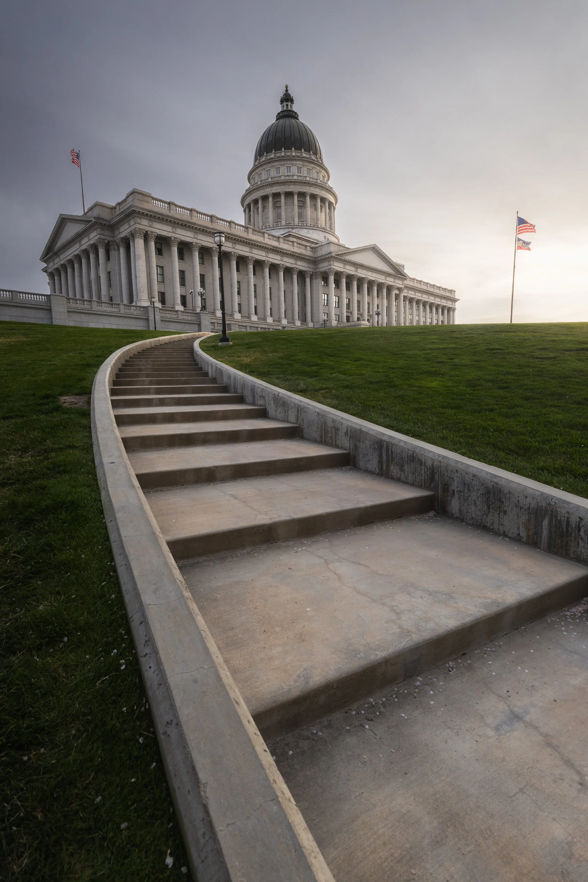 The Utah State Capitol building with wide concrete steps leading up to it, and two American flags flying on flagpoles in front of cloudy sky.