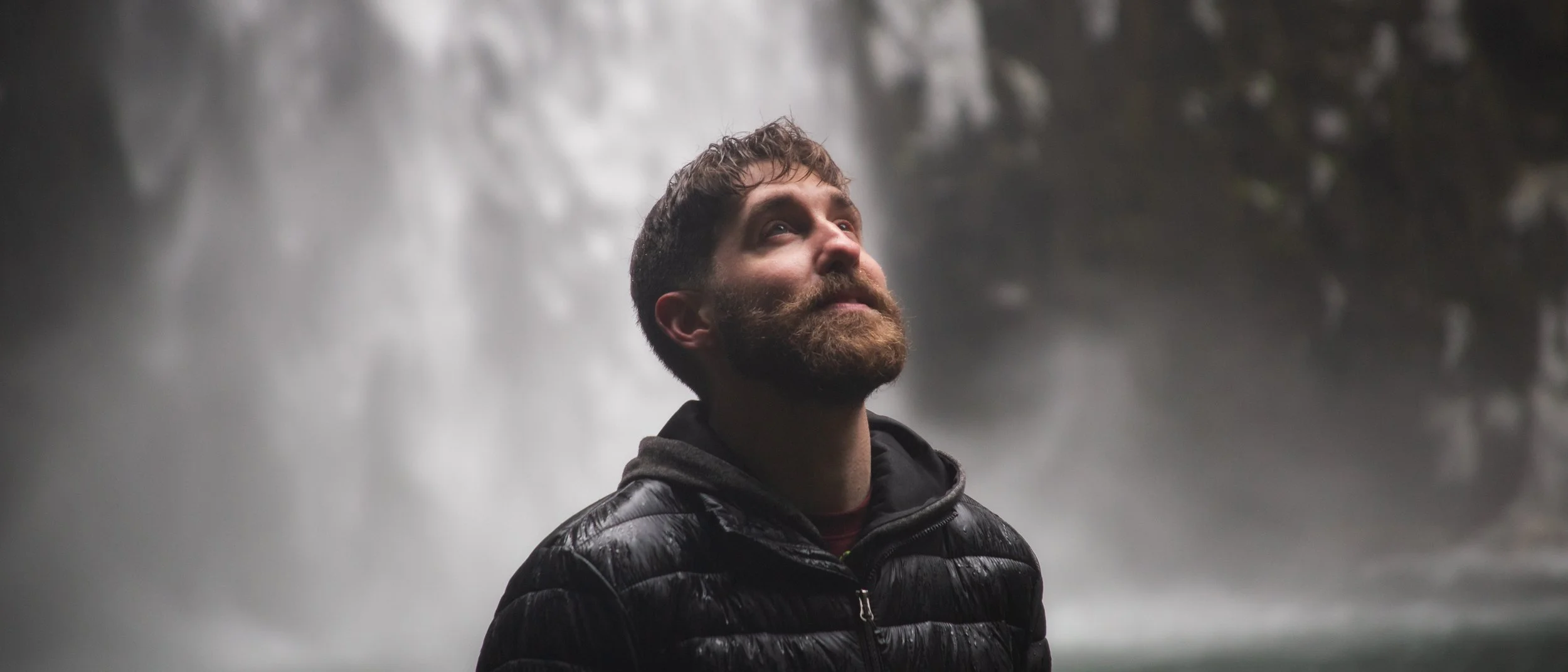 A man with a beard and short curly hair looks upward with a serene expression, wearing a black puffy jacket, standing in front of a waterfall.