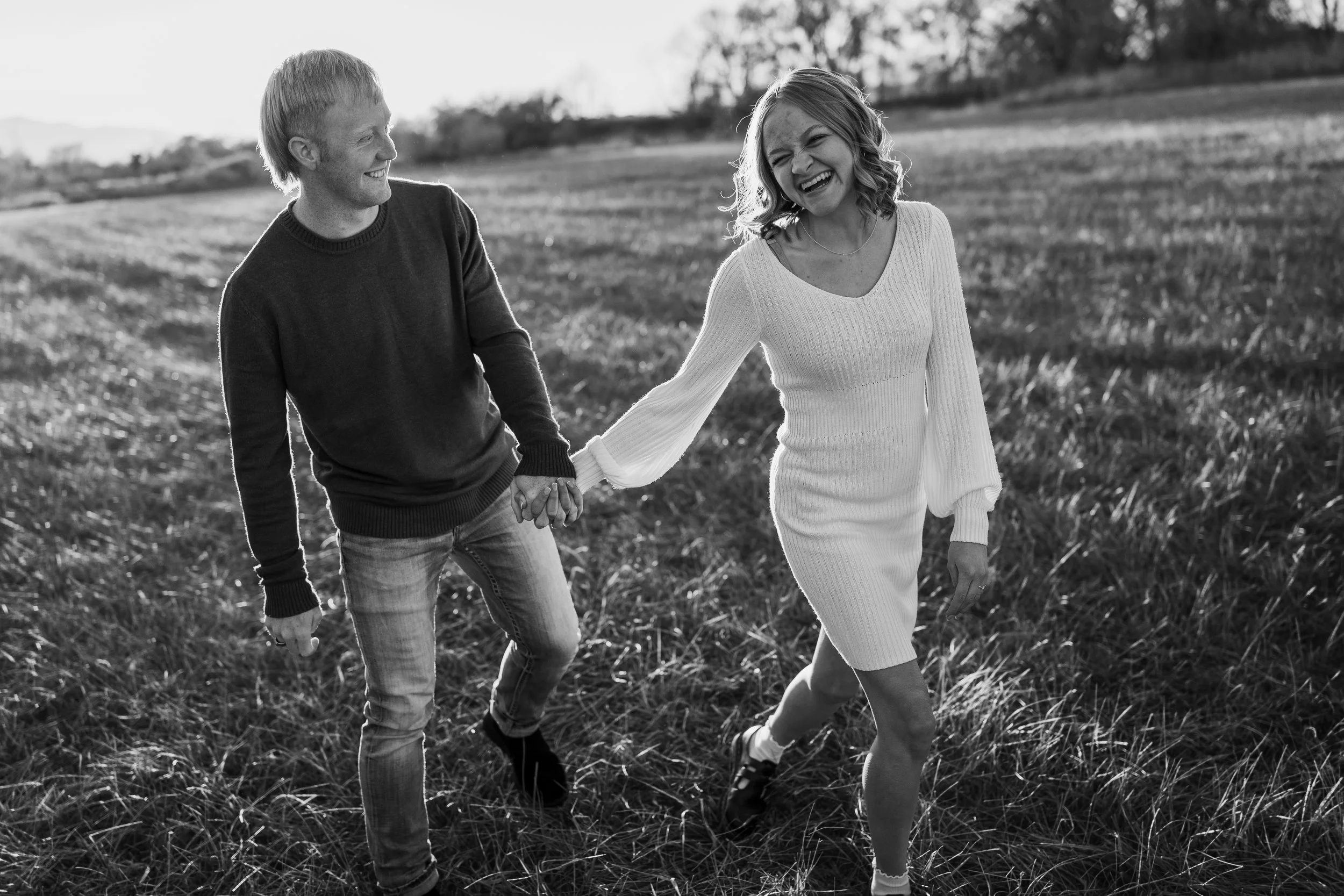 A man and woman are smiling and holding hands as they walk through a field during daylight. The woman is wearing a white dress and the man is wearing a sweater and jeans.