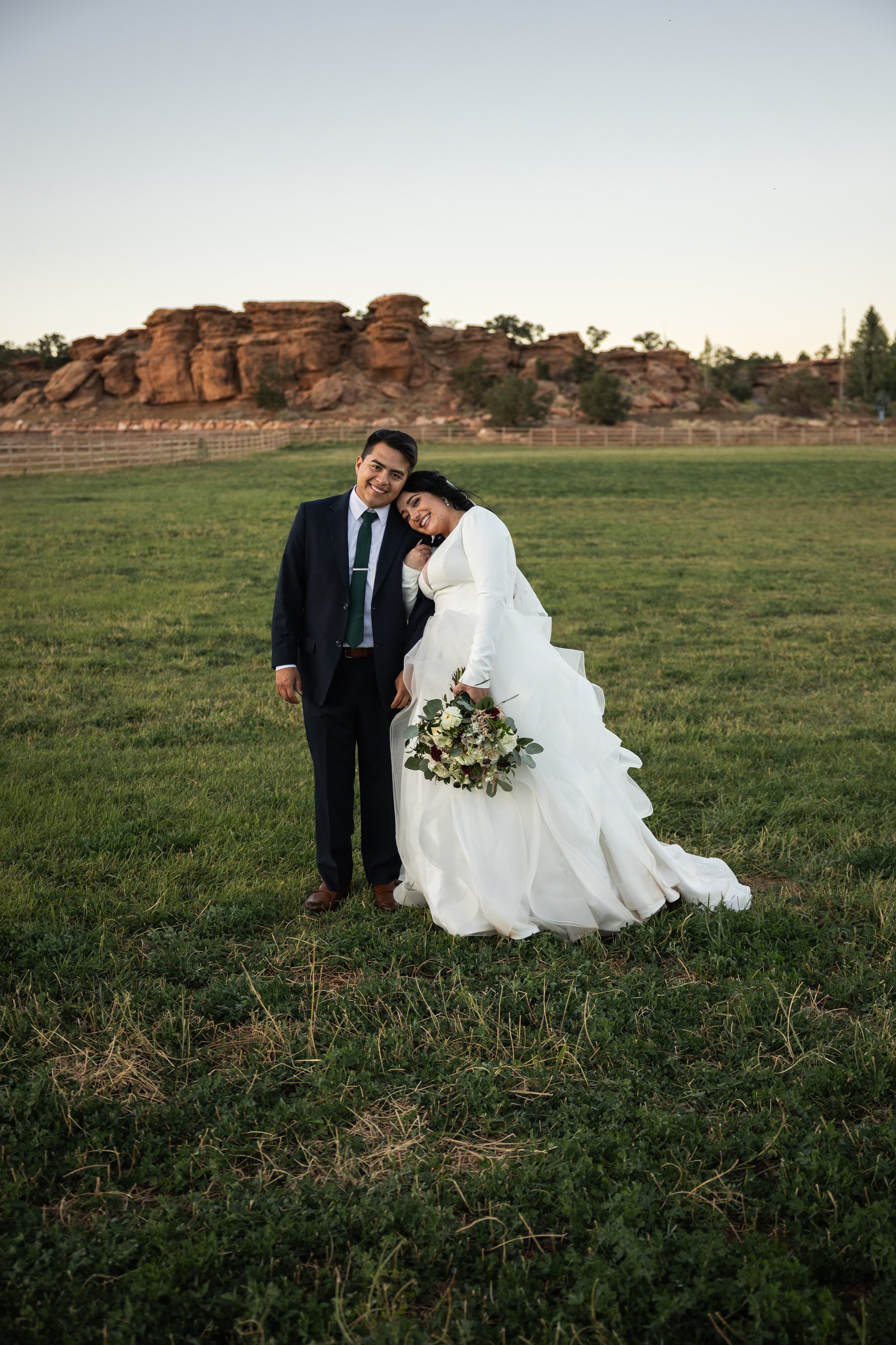 A newlywed couple standing in a grassy field at sunset, with rocky hills in the background. The woman is in a white wedding gown holding a bouquet, and she is leaning on the man's shoulder. The man is in a dark suit and green tie, smiling.
