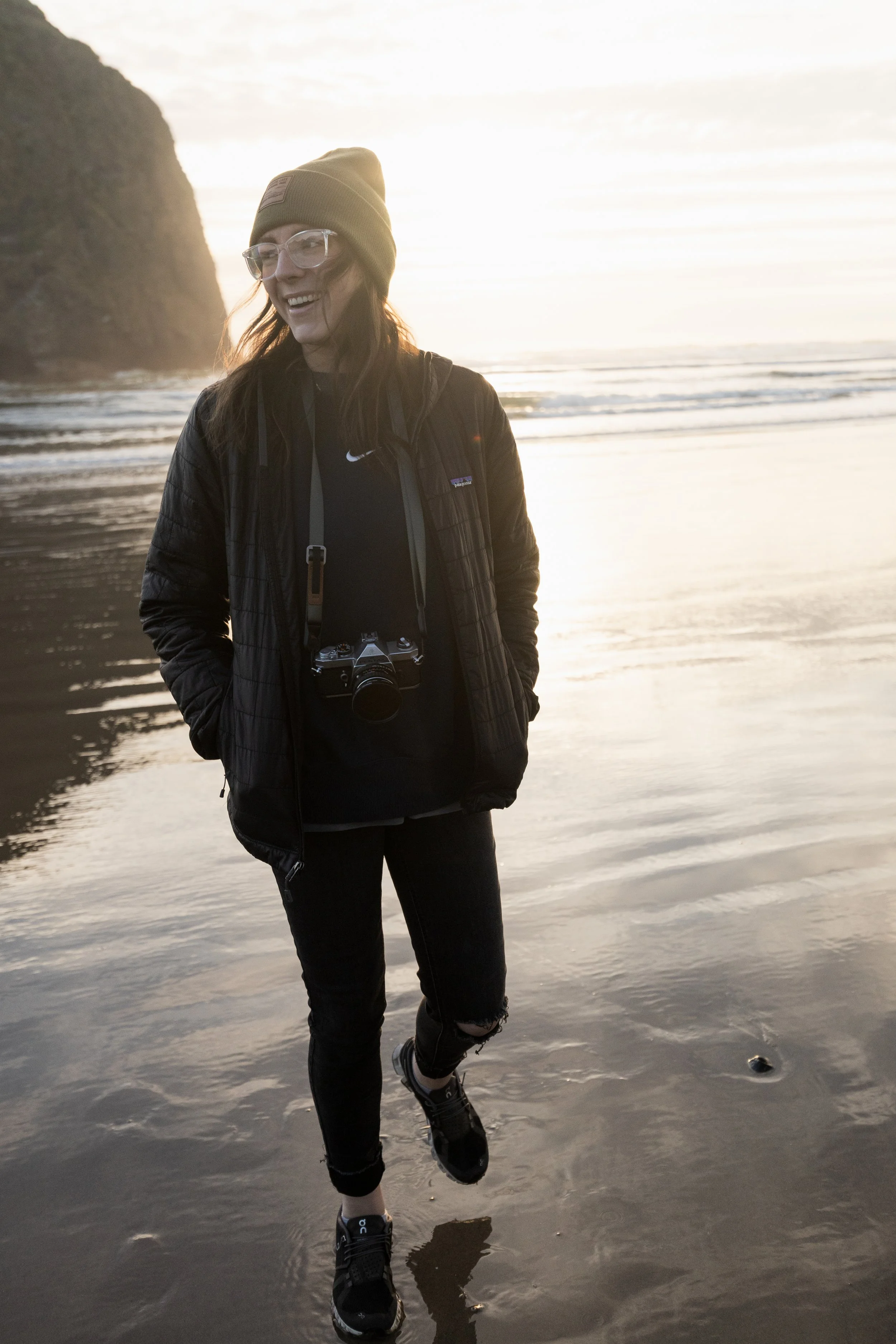 Woman smiling on the beach during sunset, wearing a beanie, glasses, a black jacket, backpack, and sneakers, with a camera hanging from her neck.