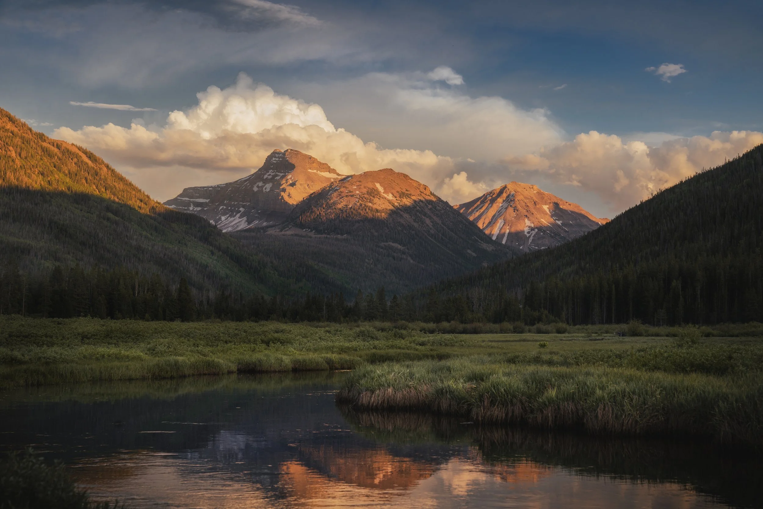 A scenic landscape of mountains with snow patches, lush green trees on the slopes, and a calm river reflecting the sky and mountains at sunset.