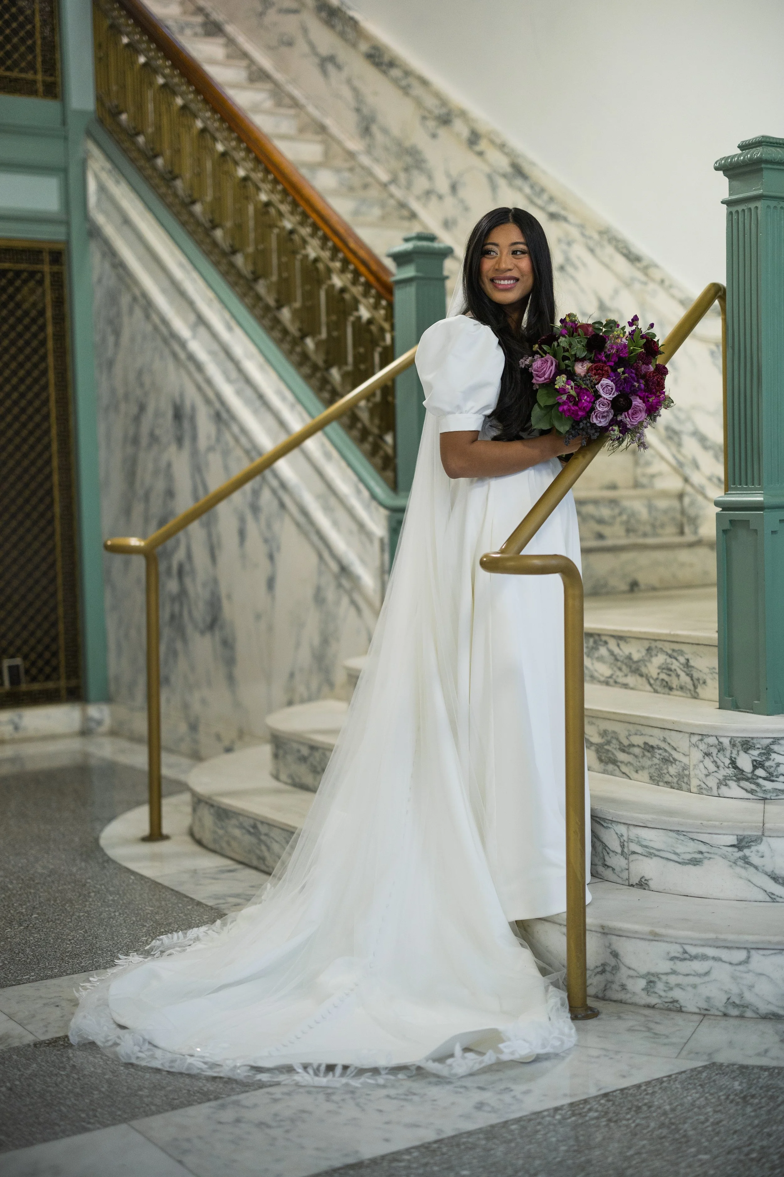 A woman wearing a white dress holding a bouquet of purple and pink flowers on a marble staircase with teal and gold railing.