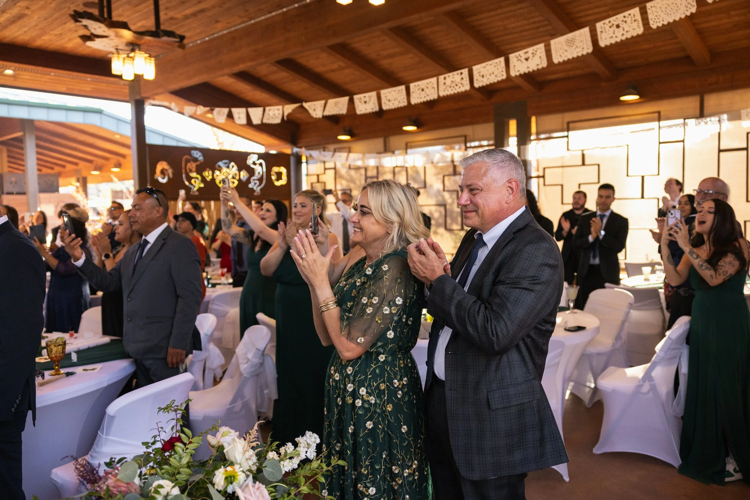 People standing and clapping at a joyful event in a decorated indoor space with wooden ceiling, some taking photos, tables with floral centerpieces.