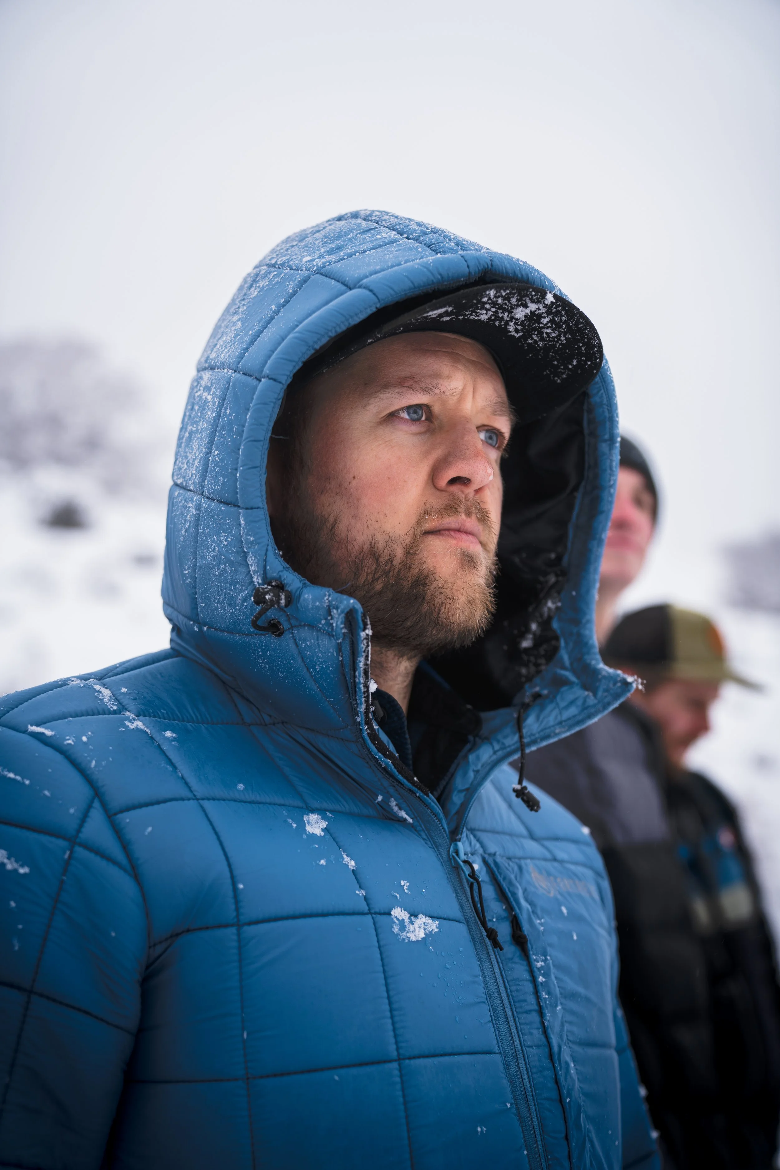 Man wearing a blue winter jacket and black cap, standing outdoors in snowy weather, with other people in the background.