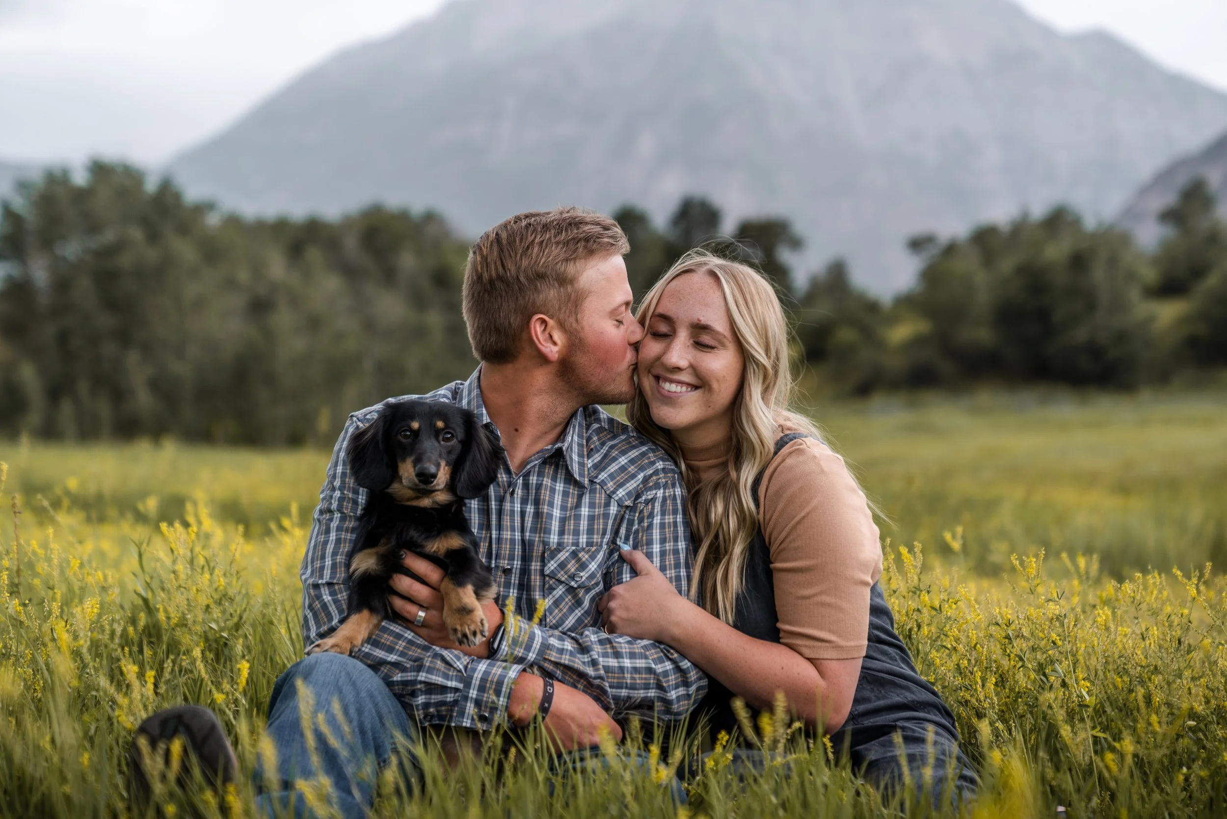 A smiling couple sitting on the grass in a field with a small dog, with mountains and trees in the background.