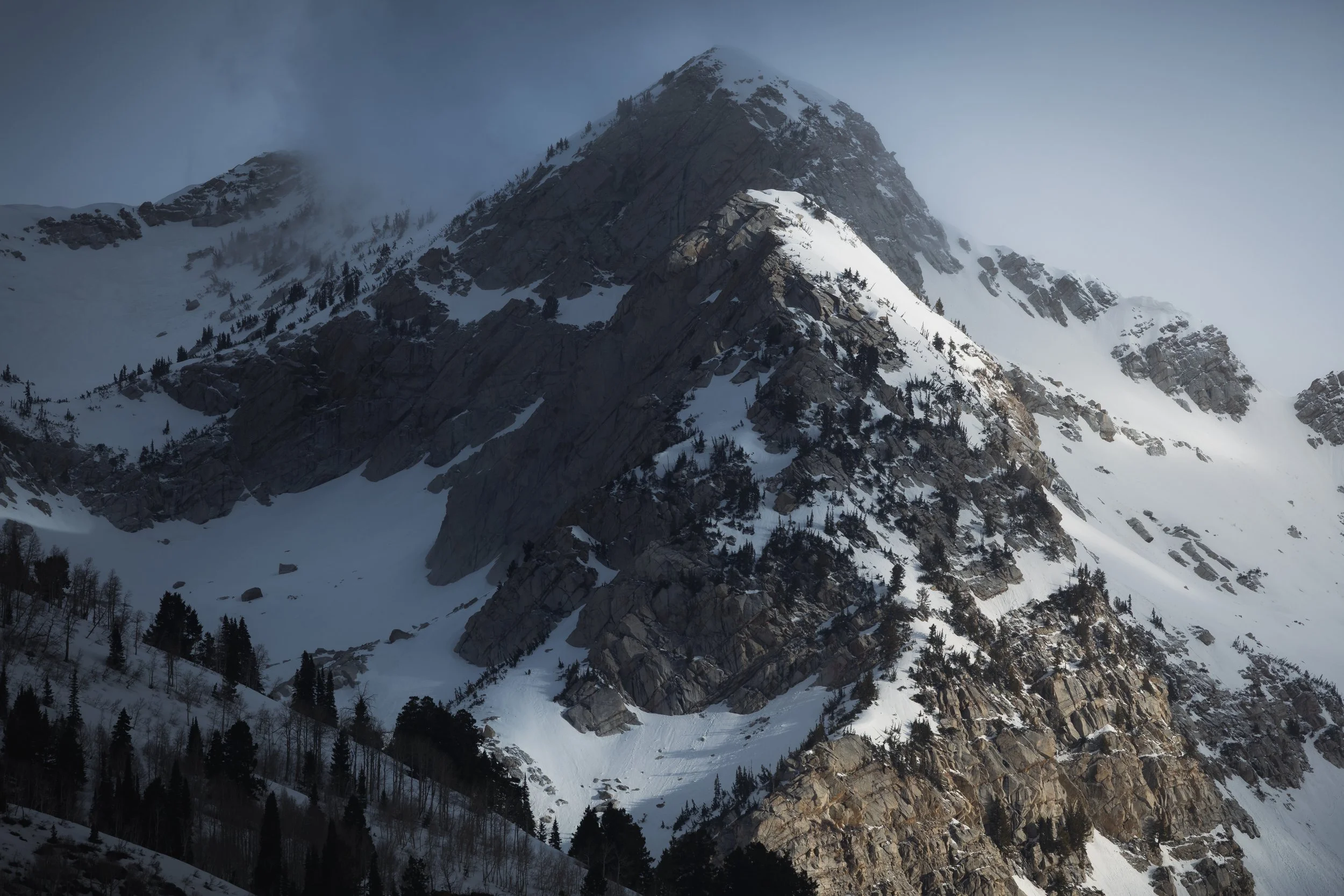 A snow-covered rugged mountain with patches of trees and clouds at the peak