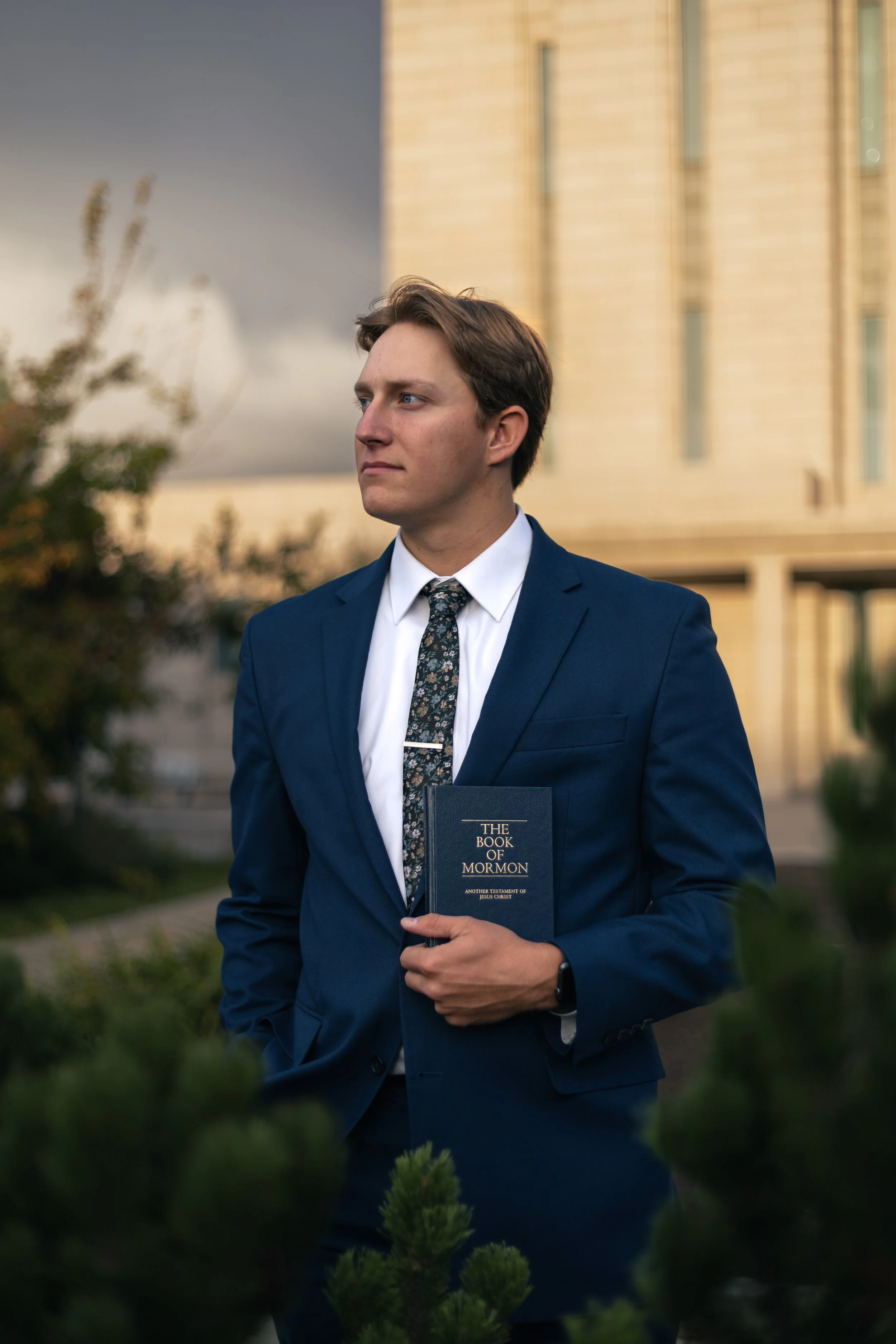 A young man in a navy suit and floral tie holding a book titled 'The Book of Mormon' outside near greenery with a tall building in the background.