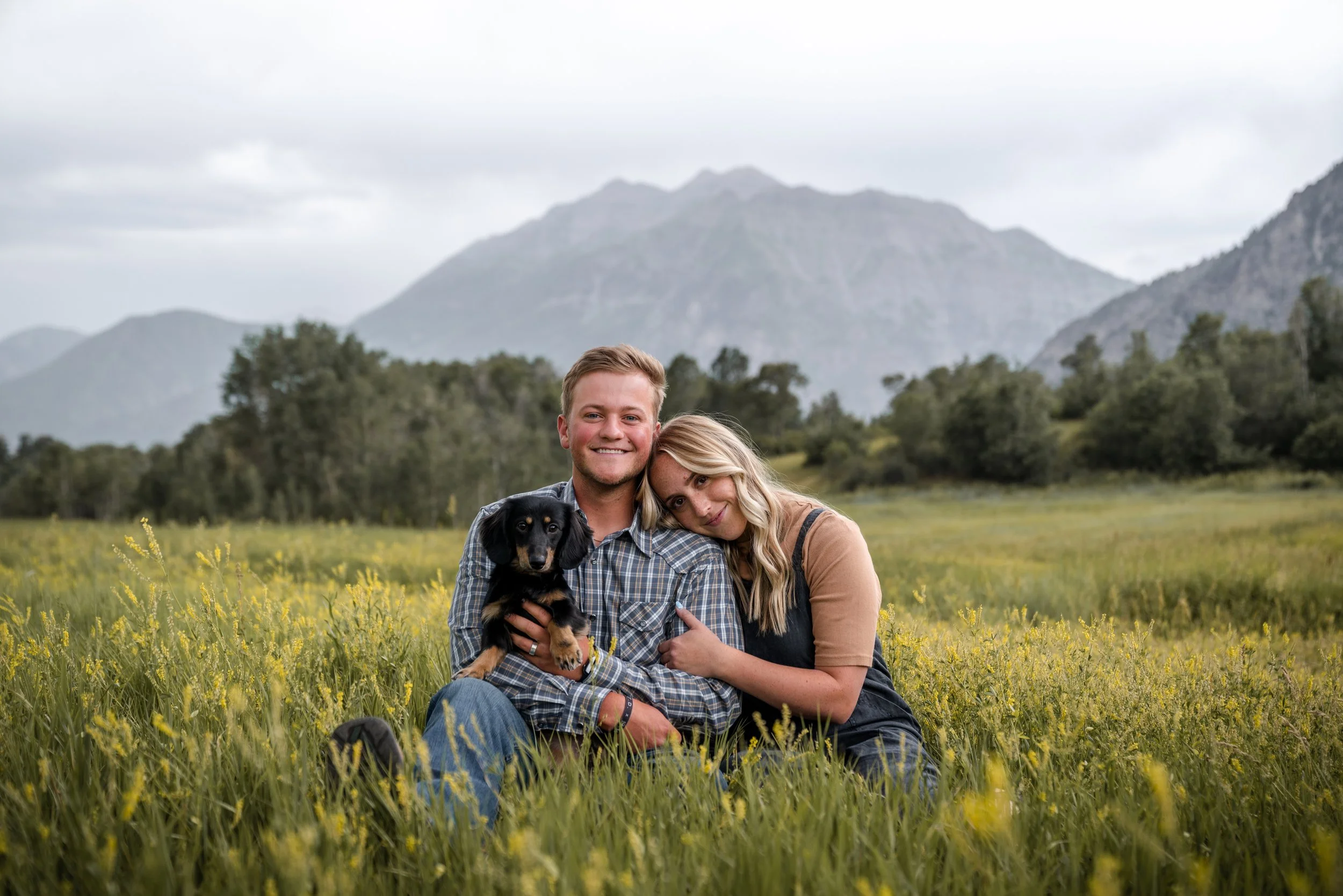 A smiling couple sitting in a field with yellow flowers, holding a small black and tan dog, with mountains and cloudy sky in the background.