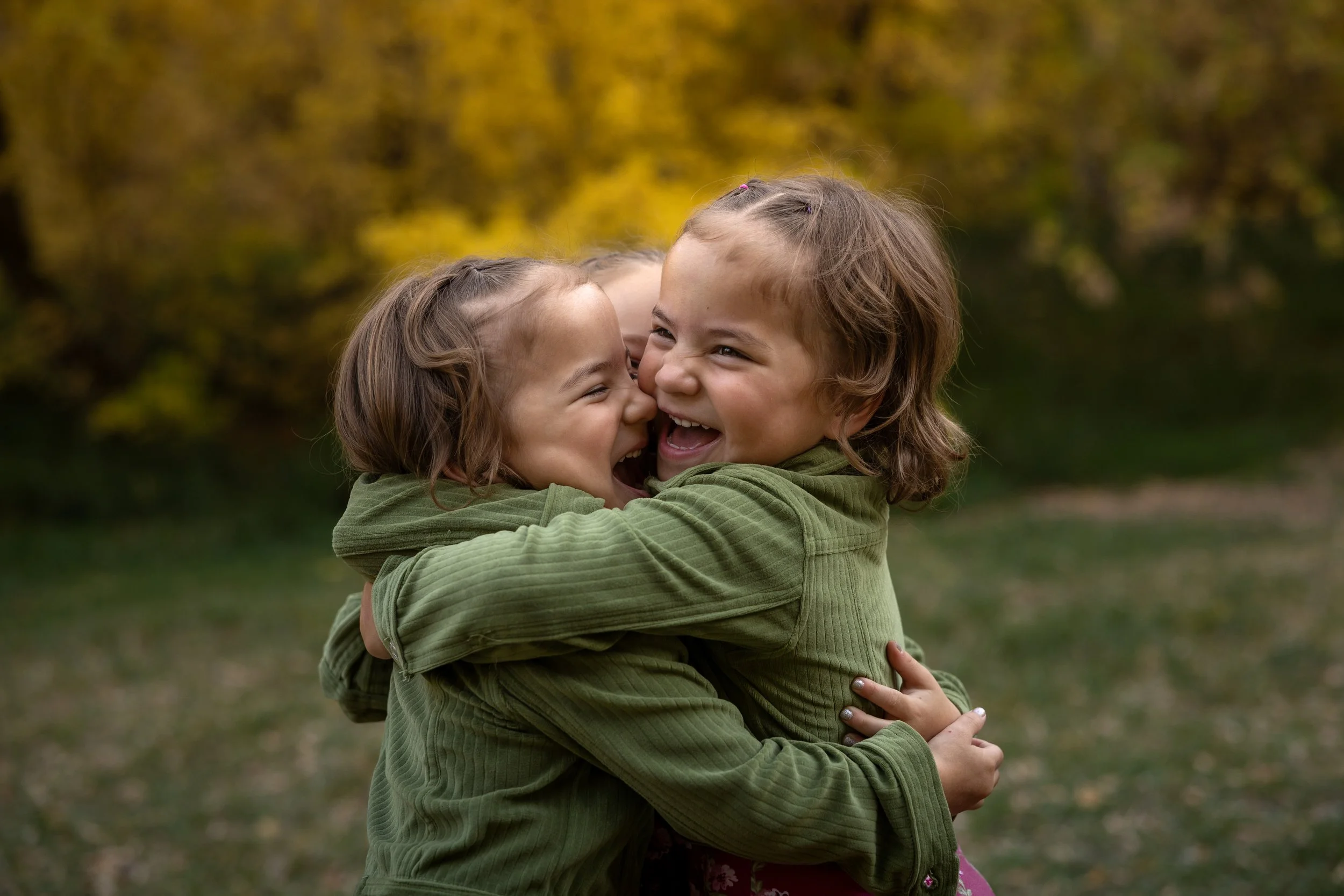 Two young girls hugging and laughing outdoors with autumn trees in the background.