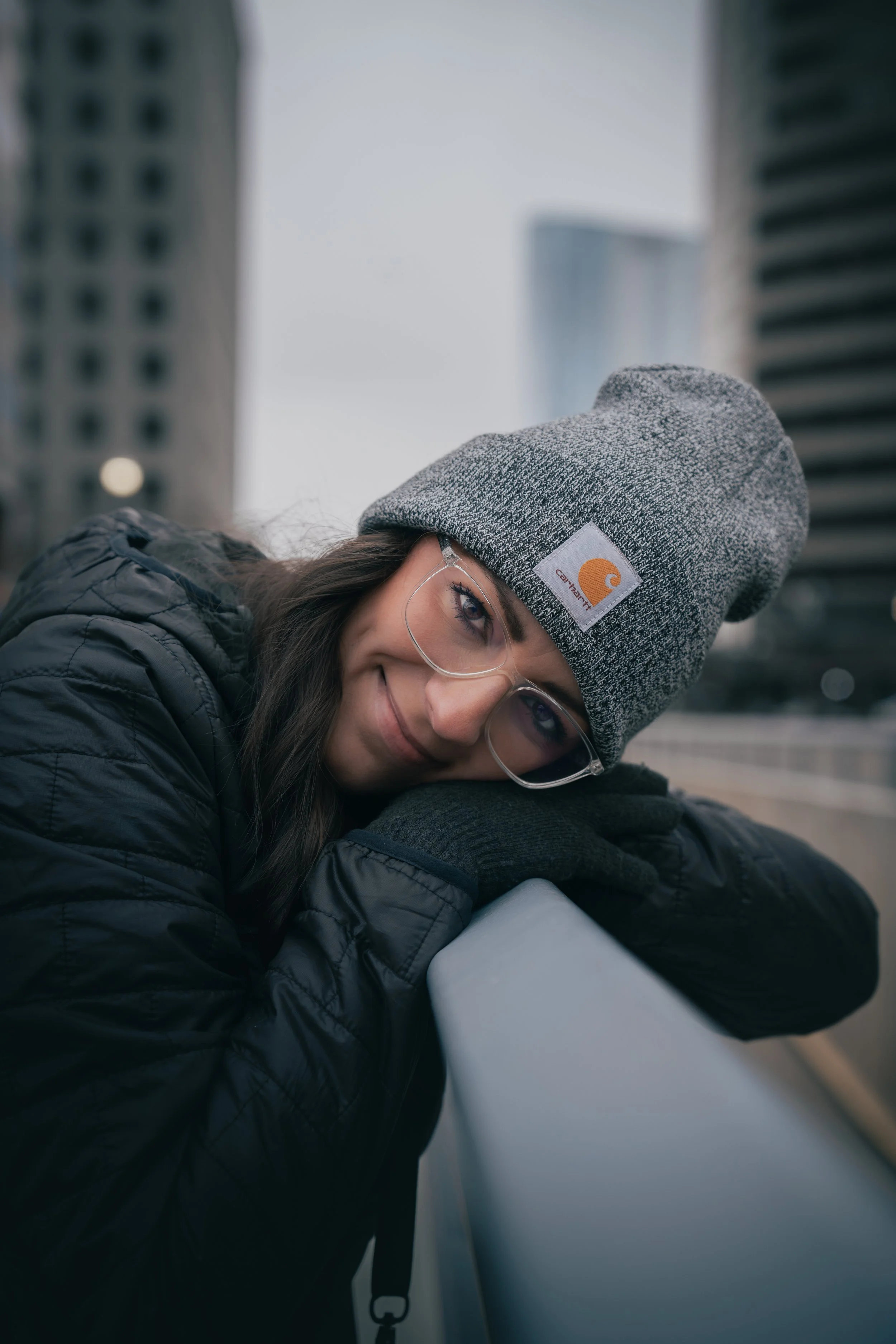 Woman in gray beanie, clear glasses, and black jacket leaning on a railing in an urban setting with tall buildings in the background.