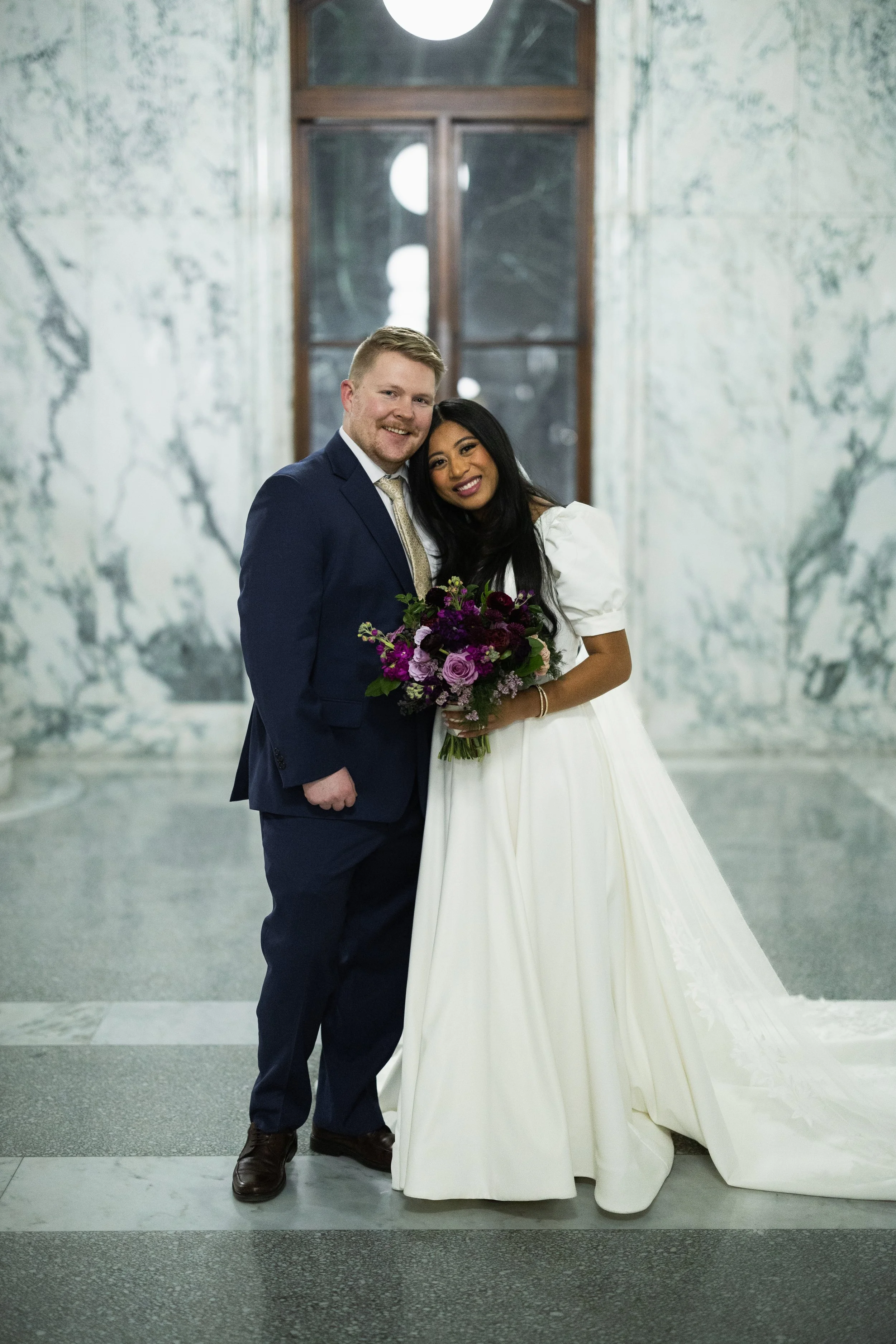 A newlywed couple poses for a photo inside a grand marble hall. The groom, dressed in a dark blue suit and tie, and the bride, wearing a white wedding gown, smile and lean close together. The bride holds a large bouquet of purple and pink flowers. Be