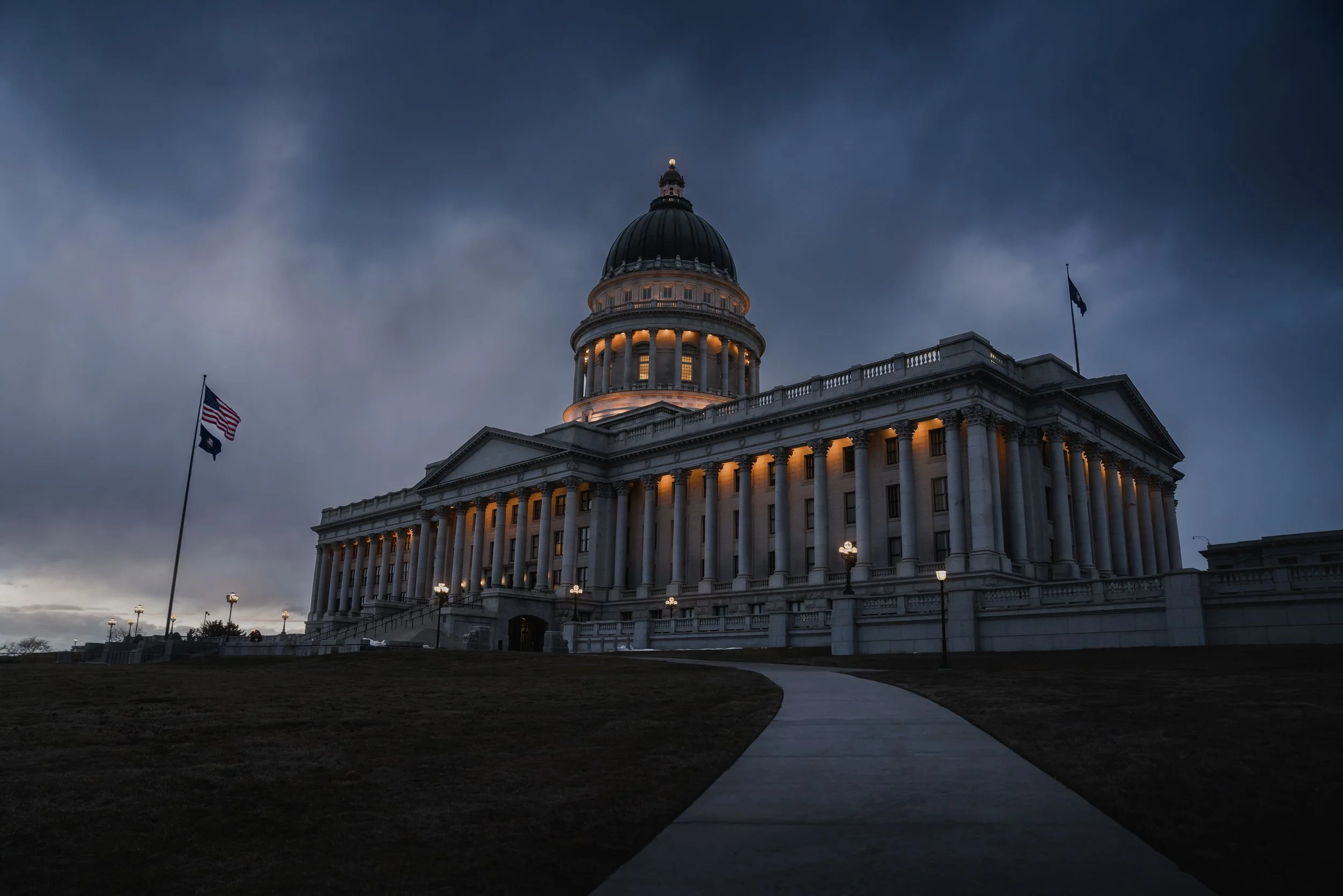 The illuminated Utah State Capitol building at dusk with dark clouds in the sky and American flags on either side.