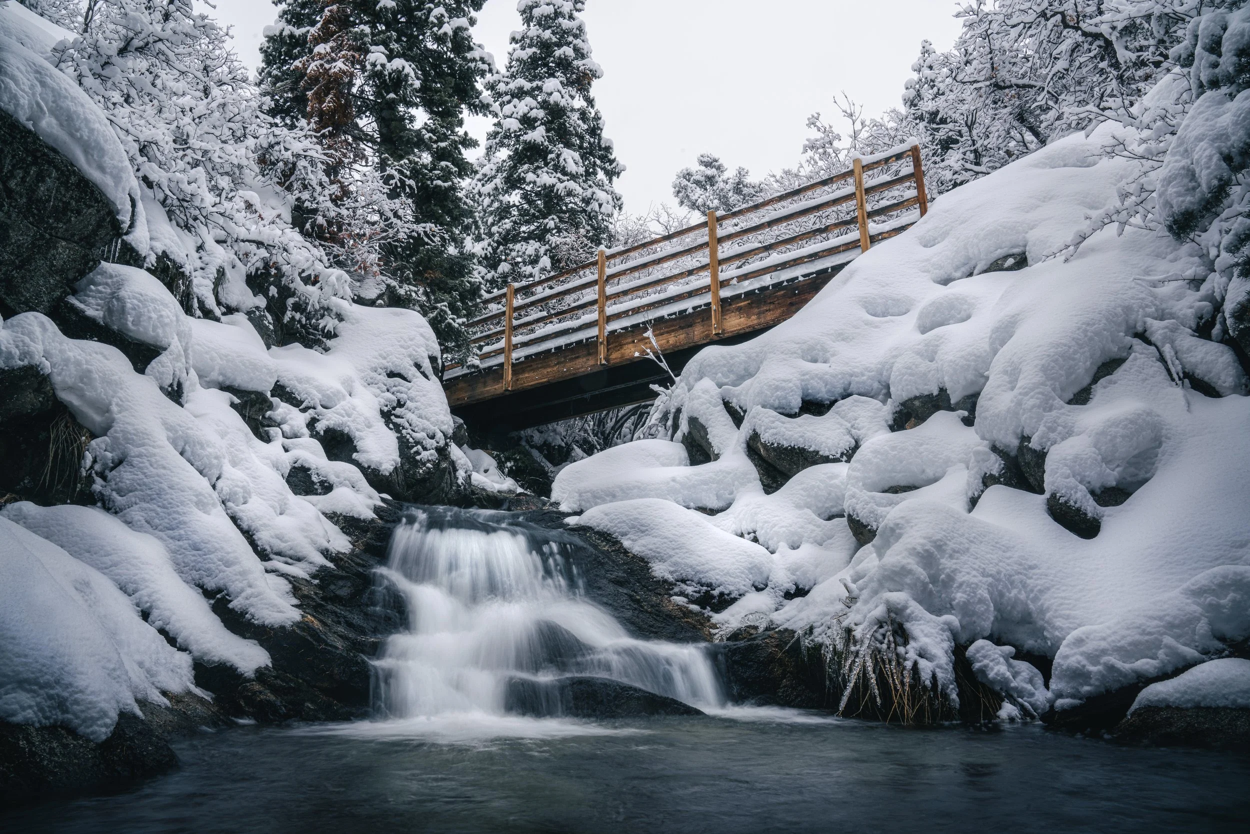 Snow-covered rocks and trees near a small waterfall with a wooden bridge crossing over the stream in a winter landscape.