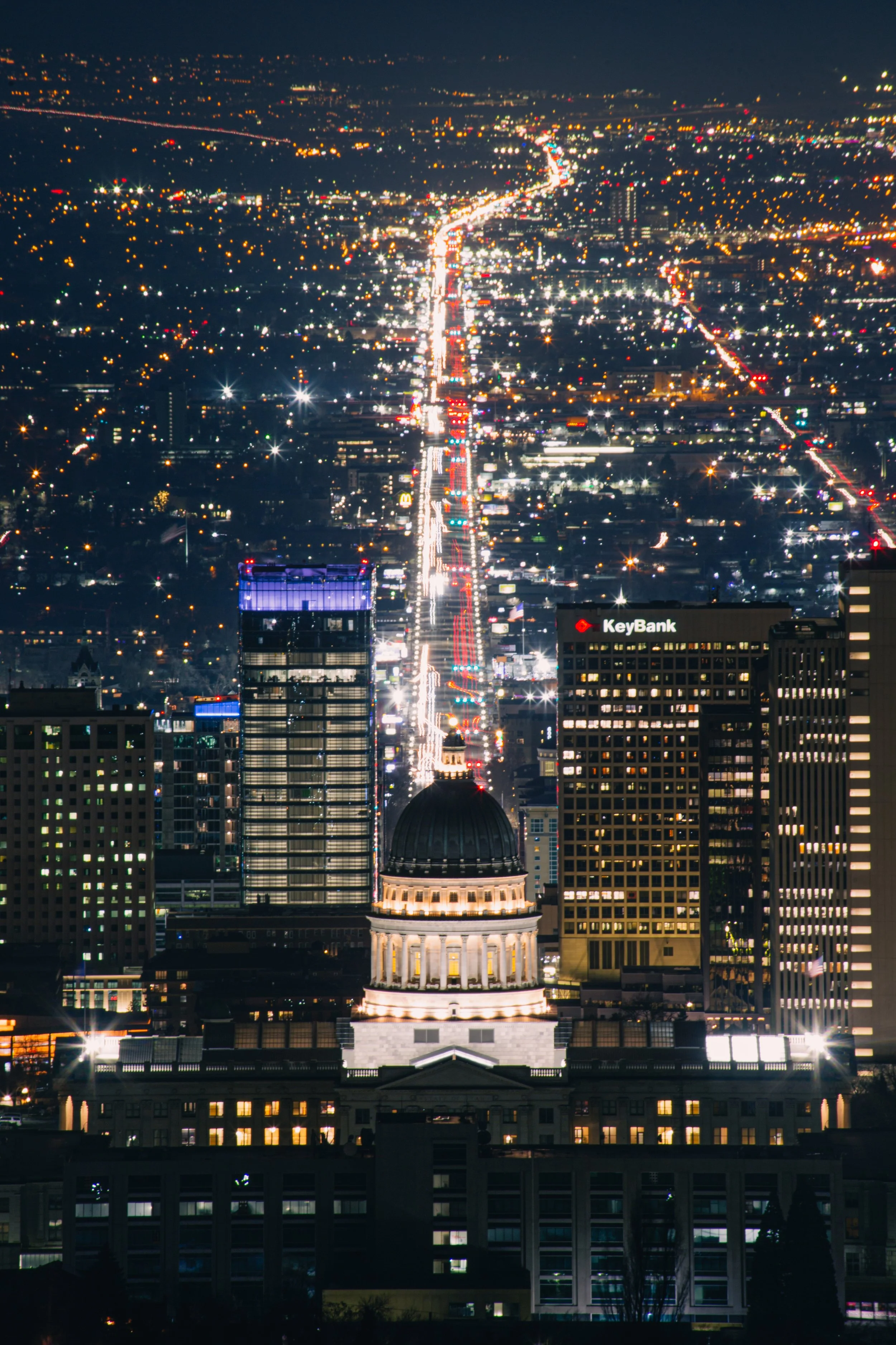 Nighttime cityscape of a downtown area with illuminated buildings and streets, featuring a historic domed building in the foreground and modern skyscrapers, including a KeyBank building, in the background.