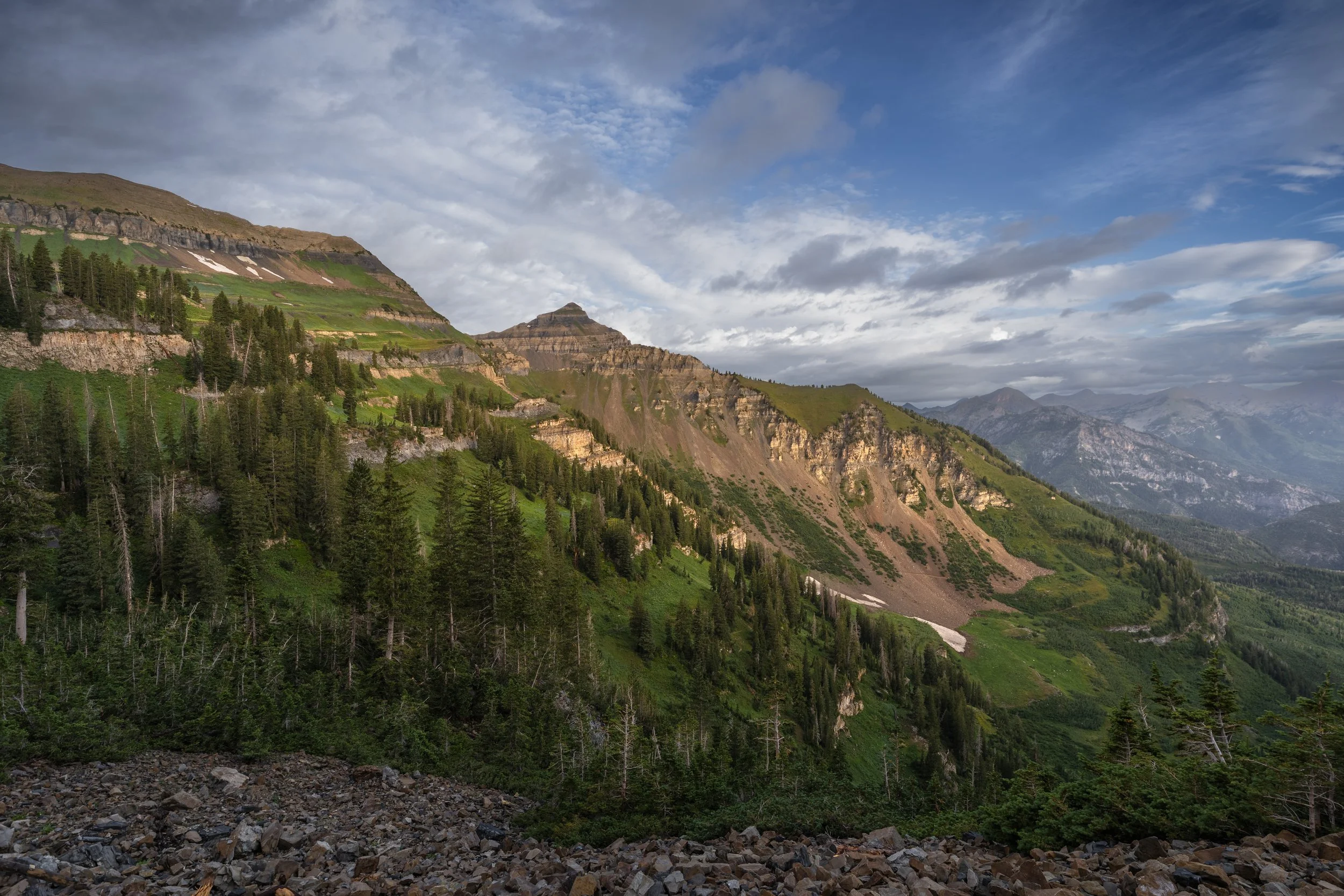 Mountain landscape with green trees, rocky slopes, and distant mountain ranges under a partly cloudy sky.