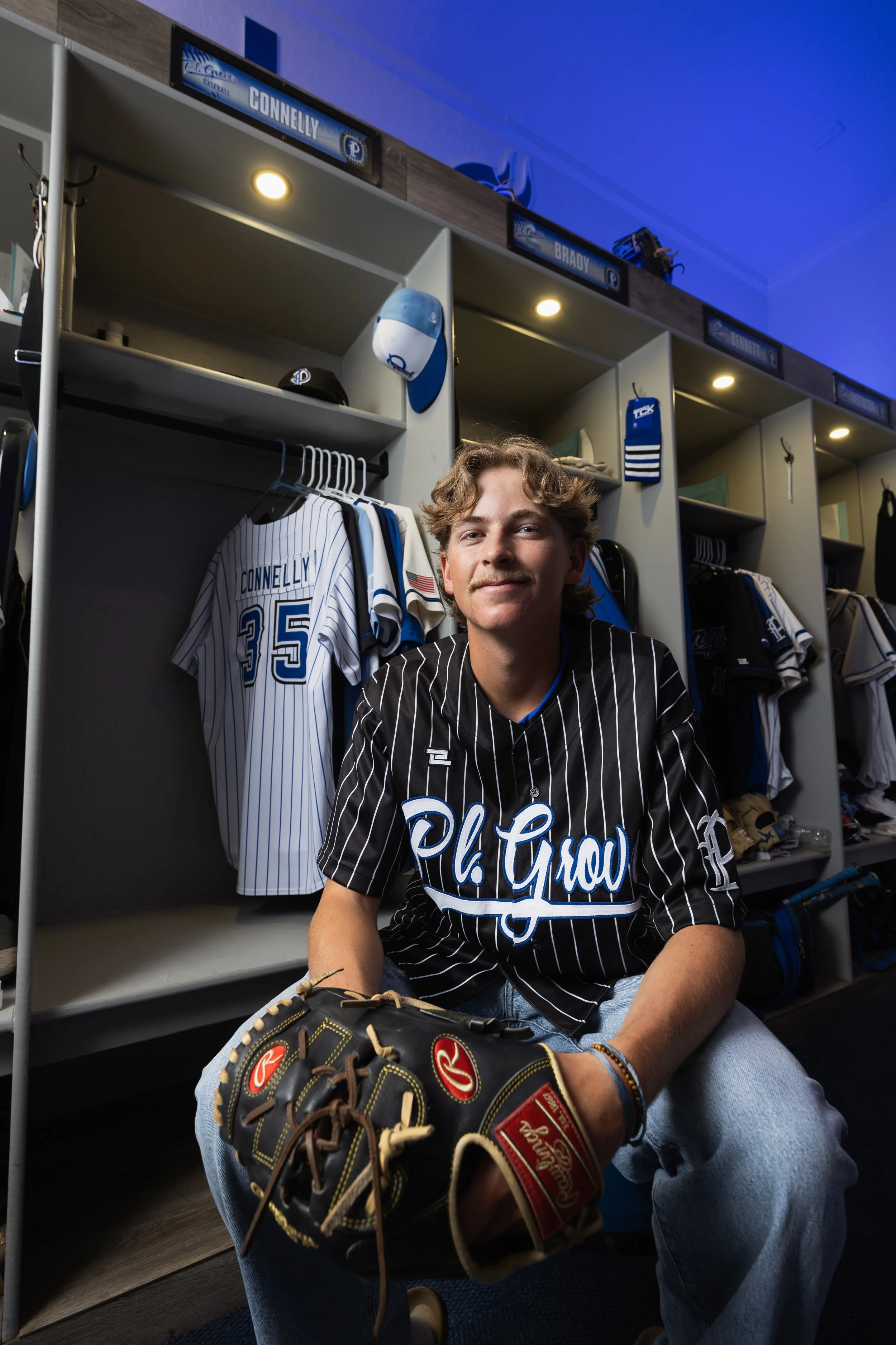 A young baseball player sitting in a locker room, holding a baseball glove, wearing a black pinstriped jersey with blue accents, surrounded by baseball equipment and jerseys.