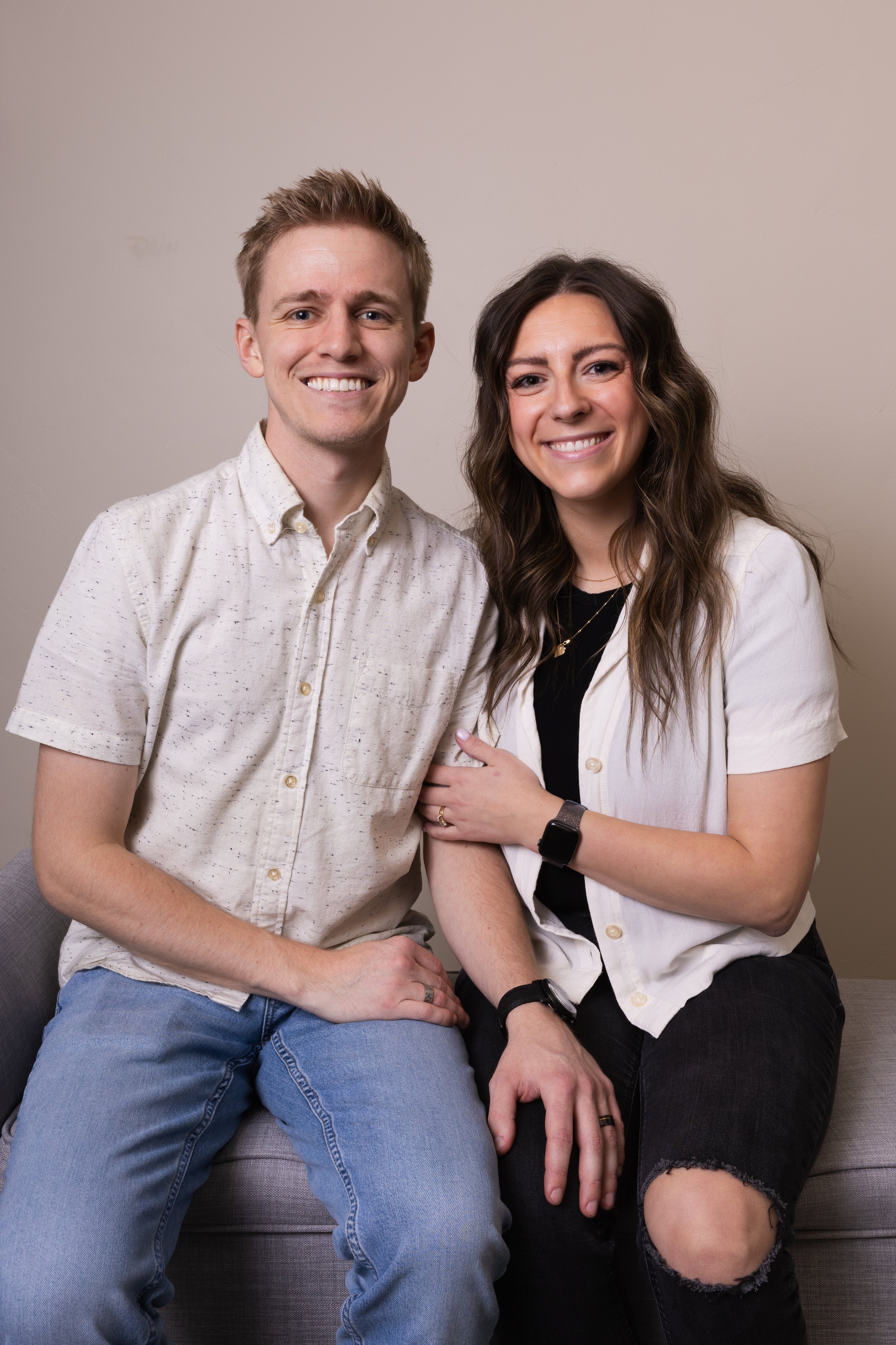 A smiling man and woman sitting close together on a gray couch, posing for a photo against a neutral background.