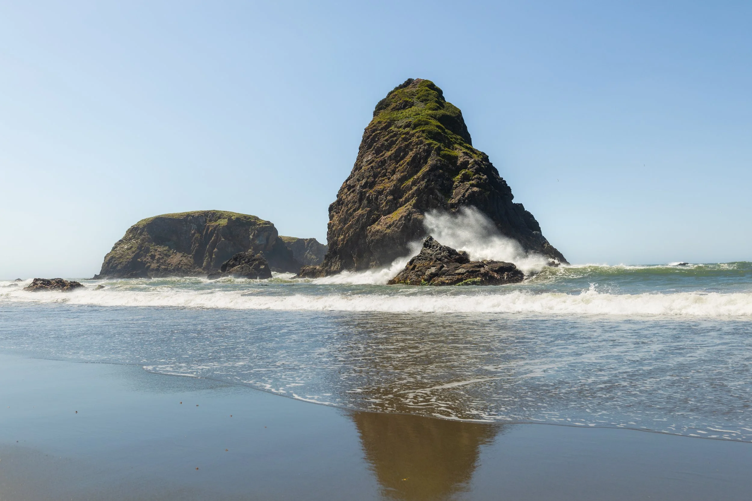 Large sea stacks with green moss, ocean waves crashing against rocks, sandy beach, clear blue sky.