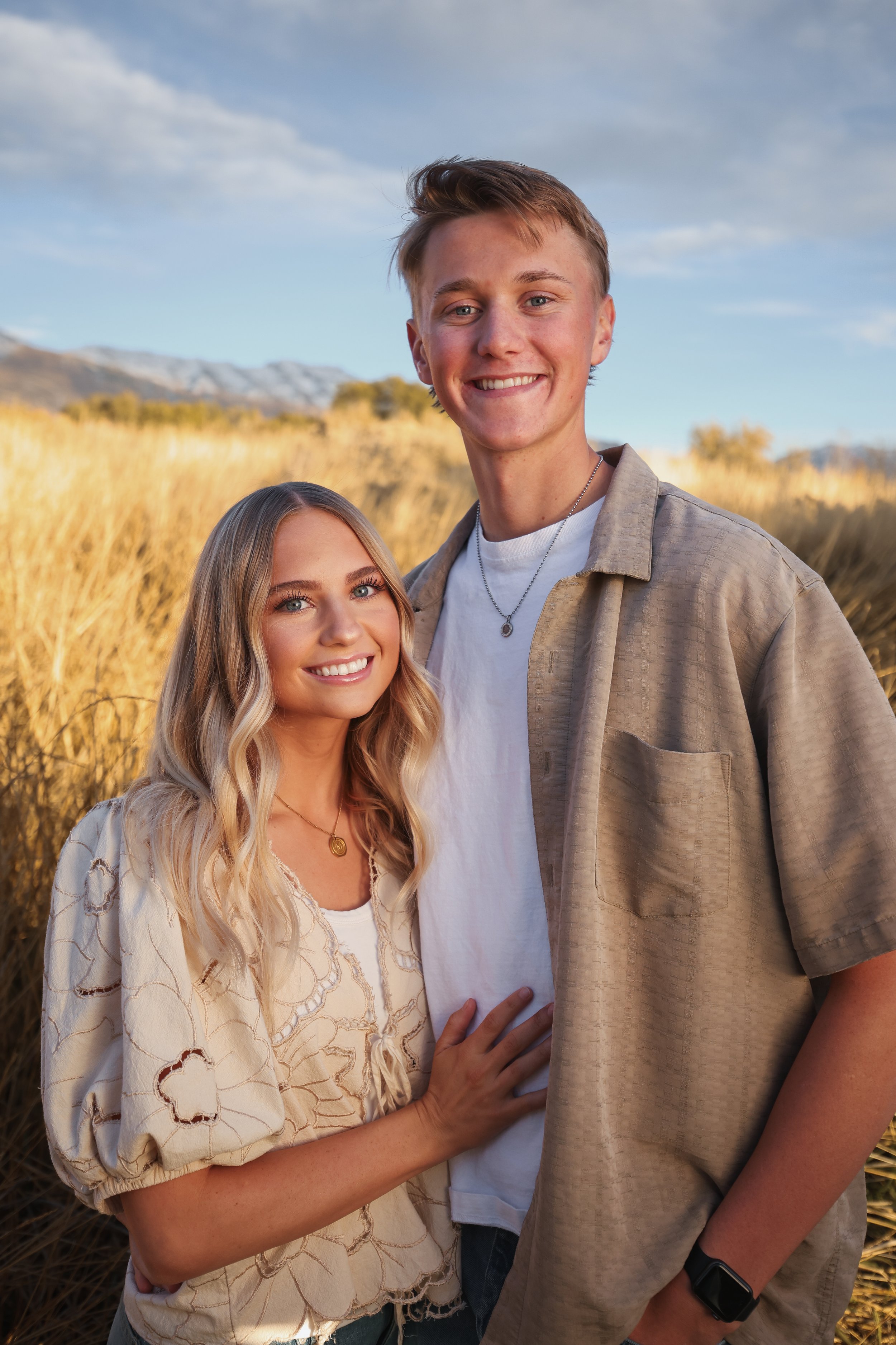 A smiling young couple standing close together outdoors in a golden field during late afternoon or early evening.