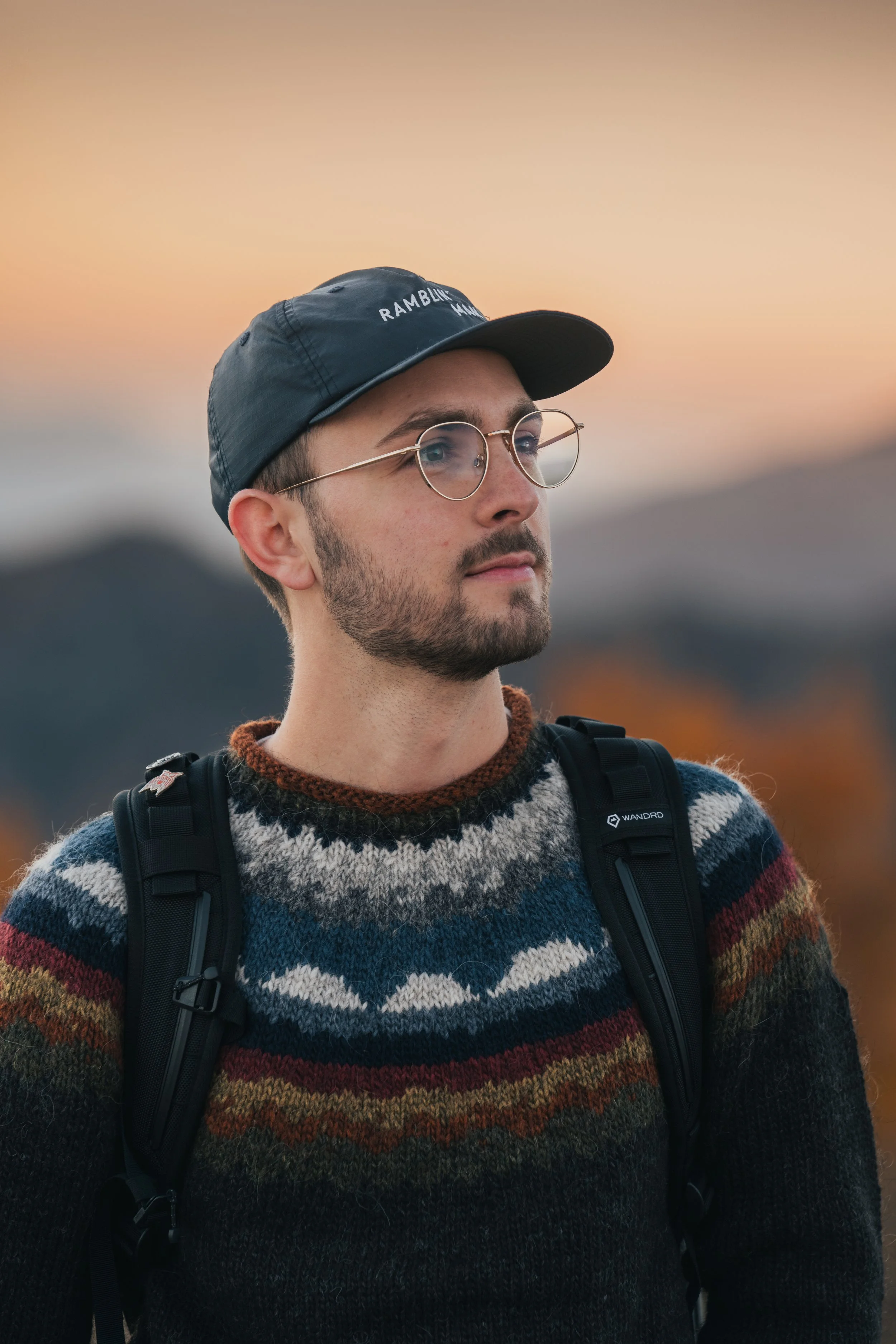 A man with glasses, a black cap, and a colorful patterned sweater outdoors at sunset, wearing a black backpack.