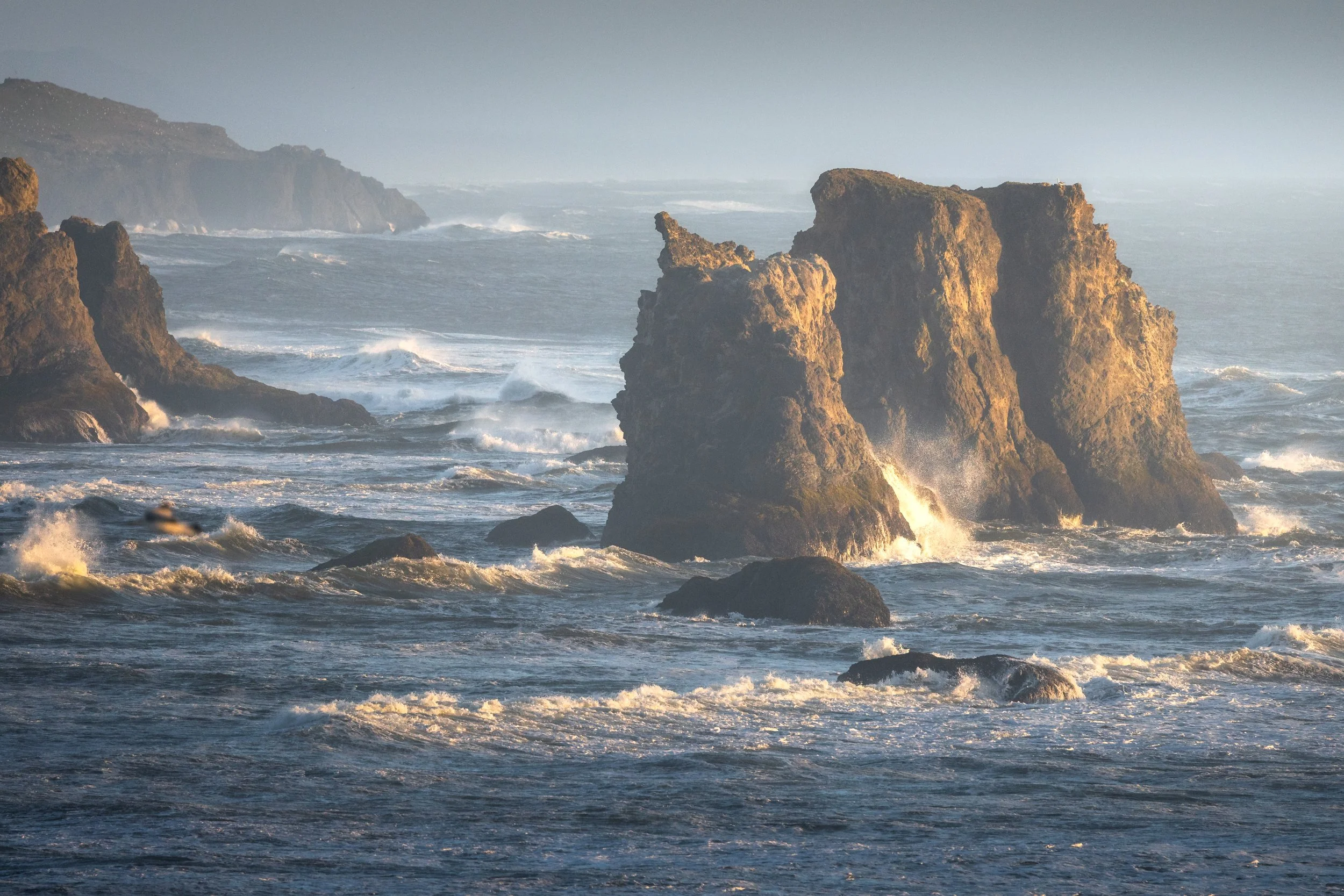 Large rock formations in the ocean with waves crashing against them, under a hazy sky.