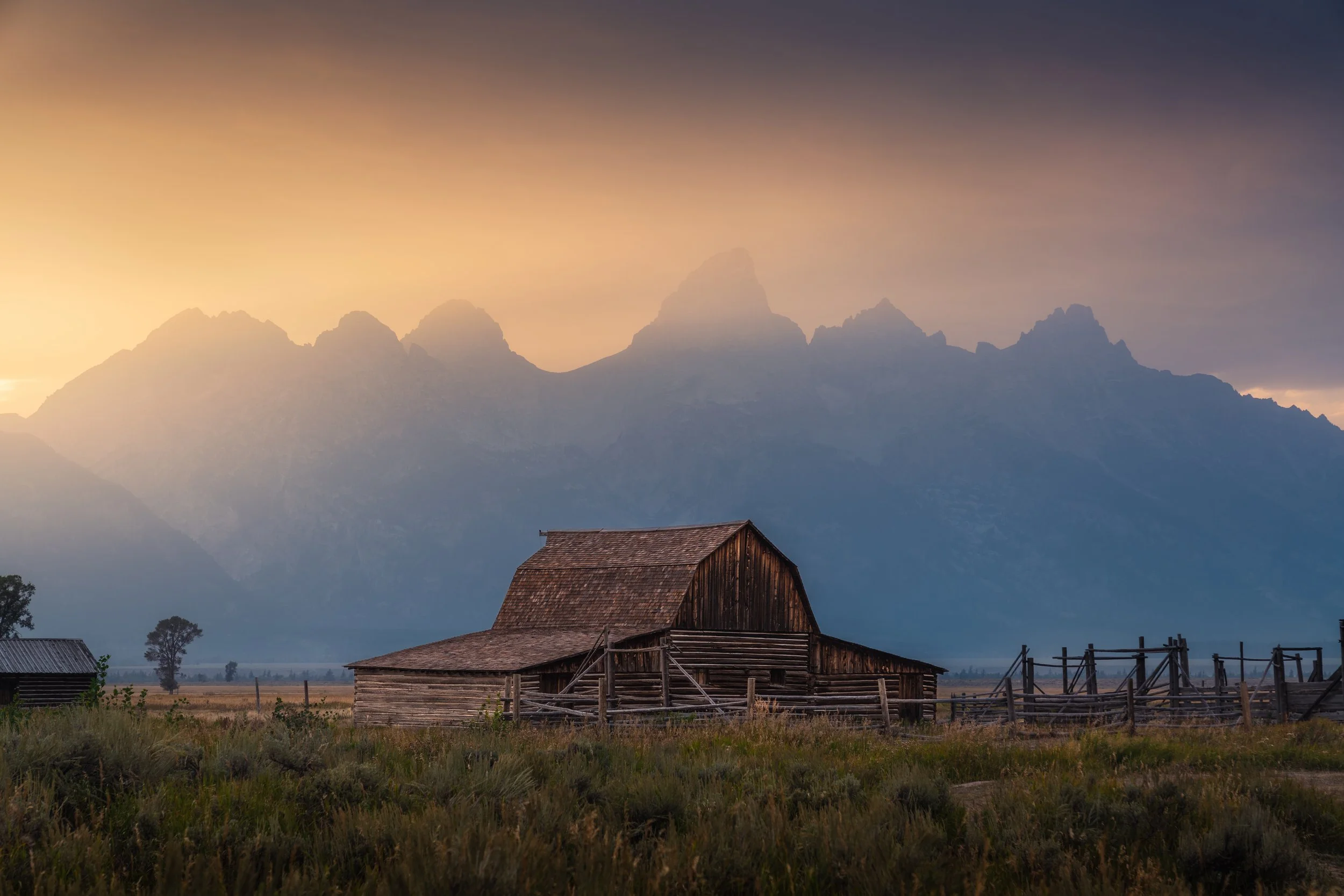 An old wooden barn in a grassy field with mountains in the background under a cloudy sky at sunset