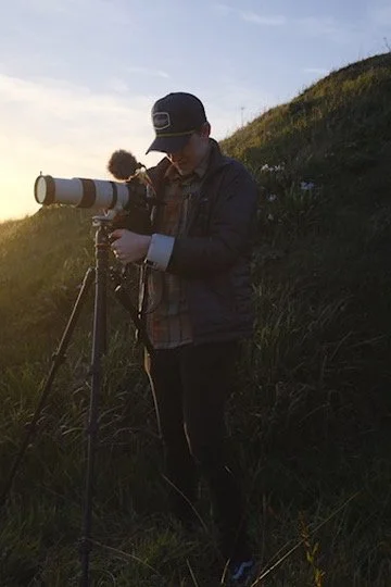 A person wearing a cap and jacket setting up a camera on a tripod during sunset in a grassy outdoor area.